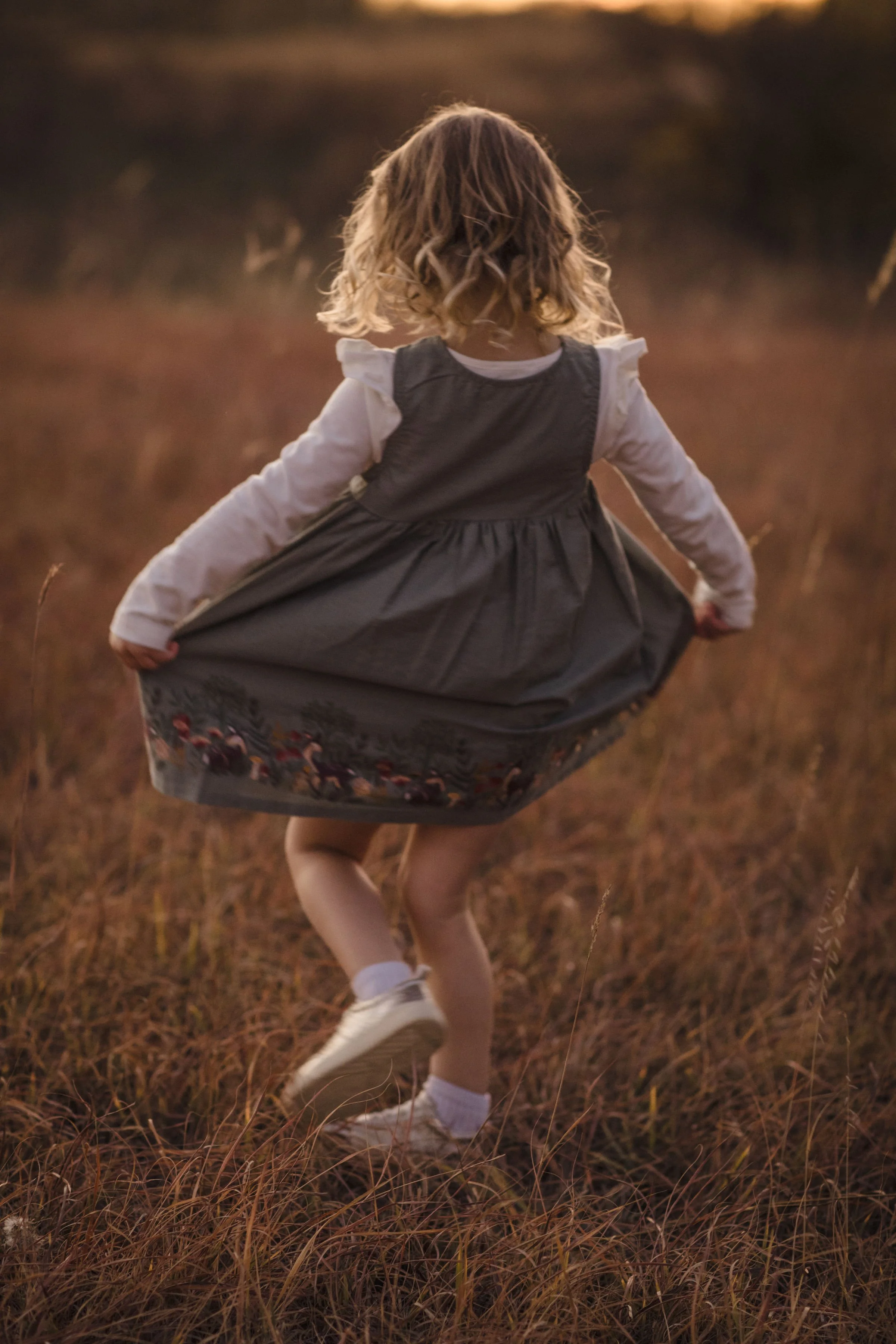 A young girl with curly blonde hair running through a field of tall, dry grass during sunset, wearing a gray dress with a printed hem, white long-sleeve shirt, and sneakers.
