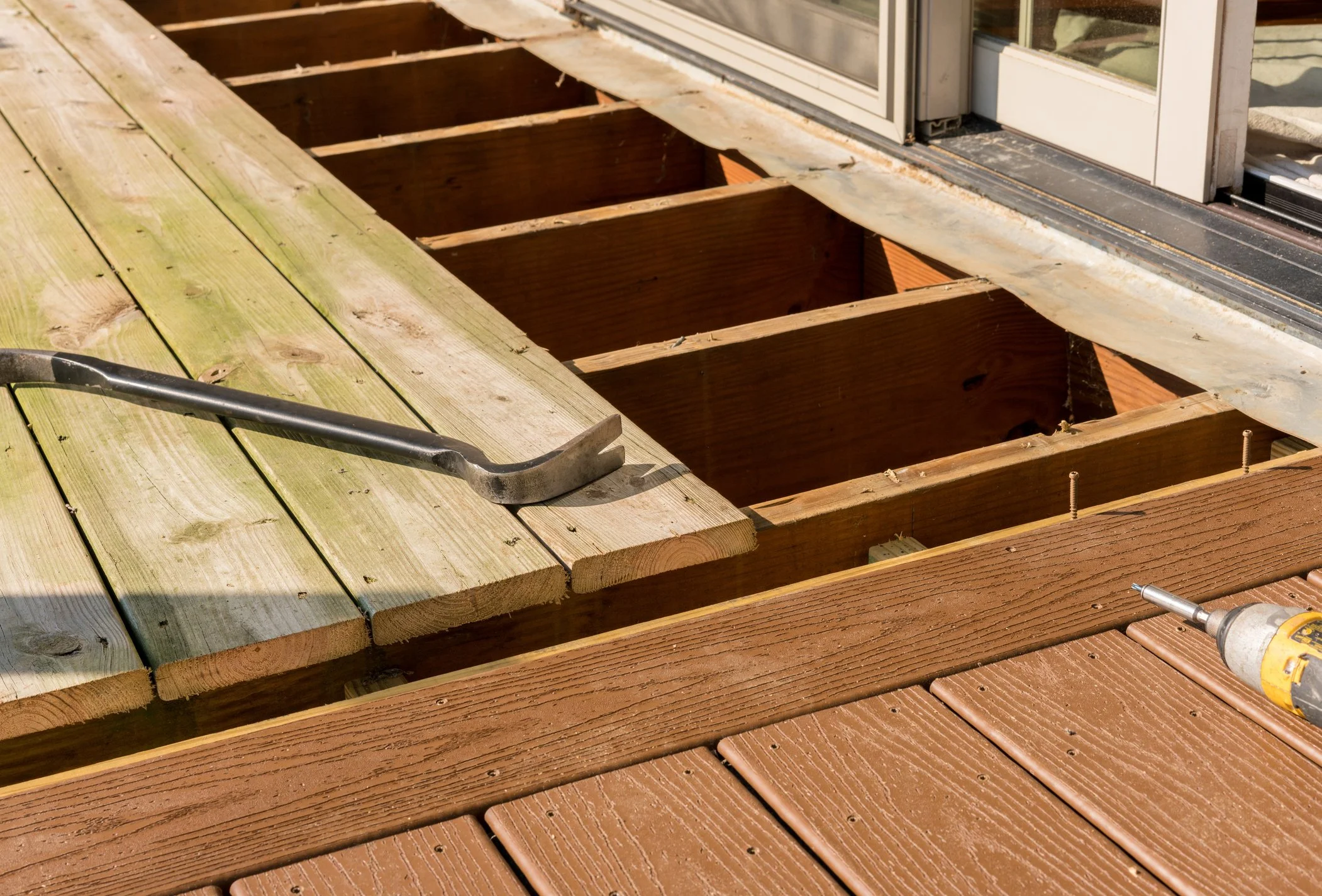 Close-up of a wooden deck under construction with some planks removed, showing the underlying framework, a crowbar resting on the unfinished deck, and a cordless drill nearby.