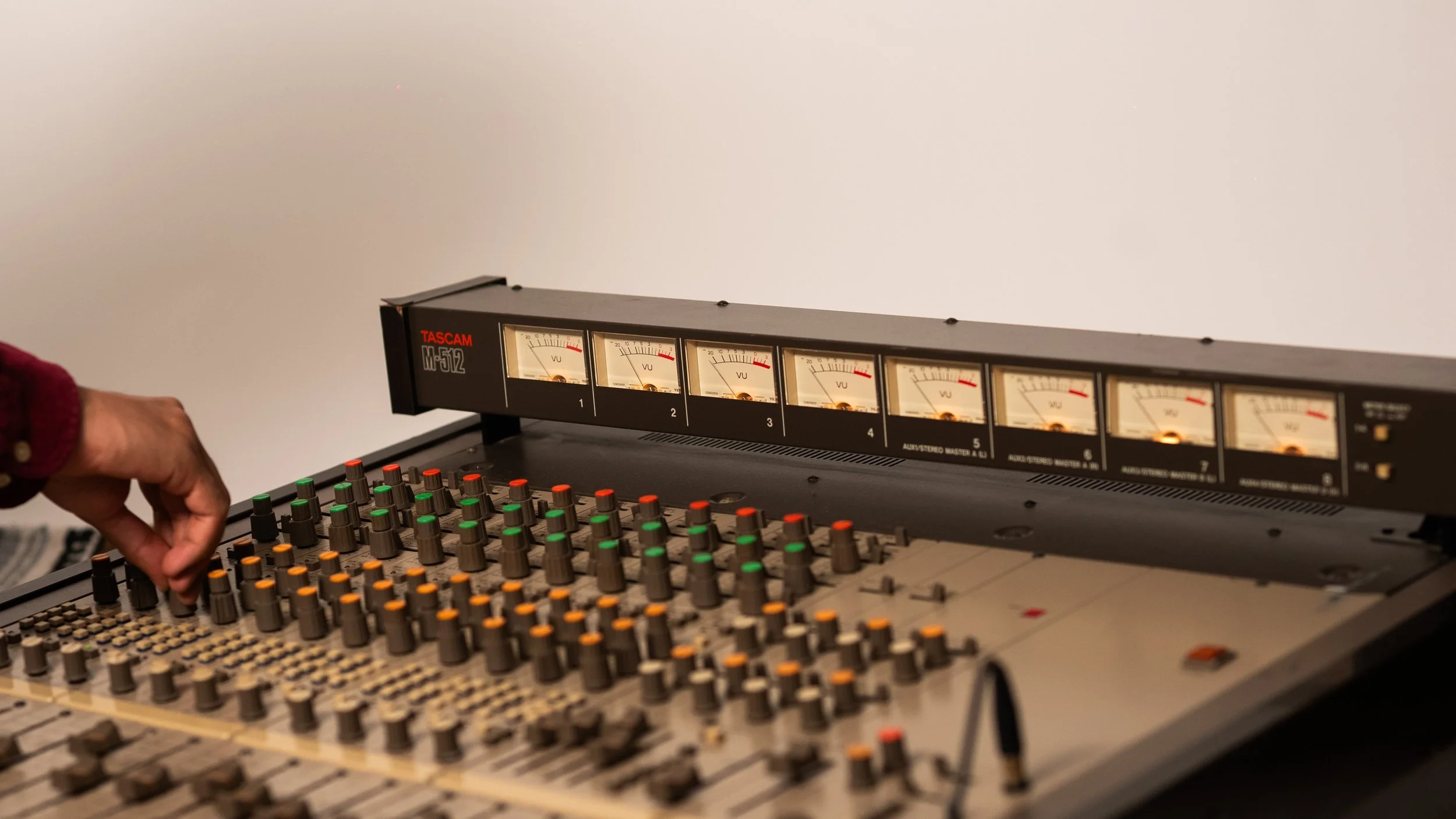 A person adjusting volume knobs on a professional audio mixing console with a VU meter display above.