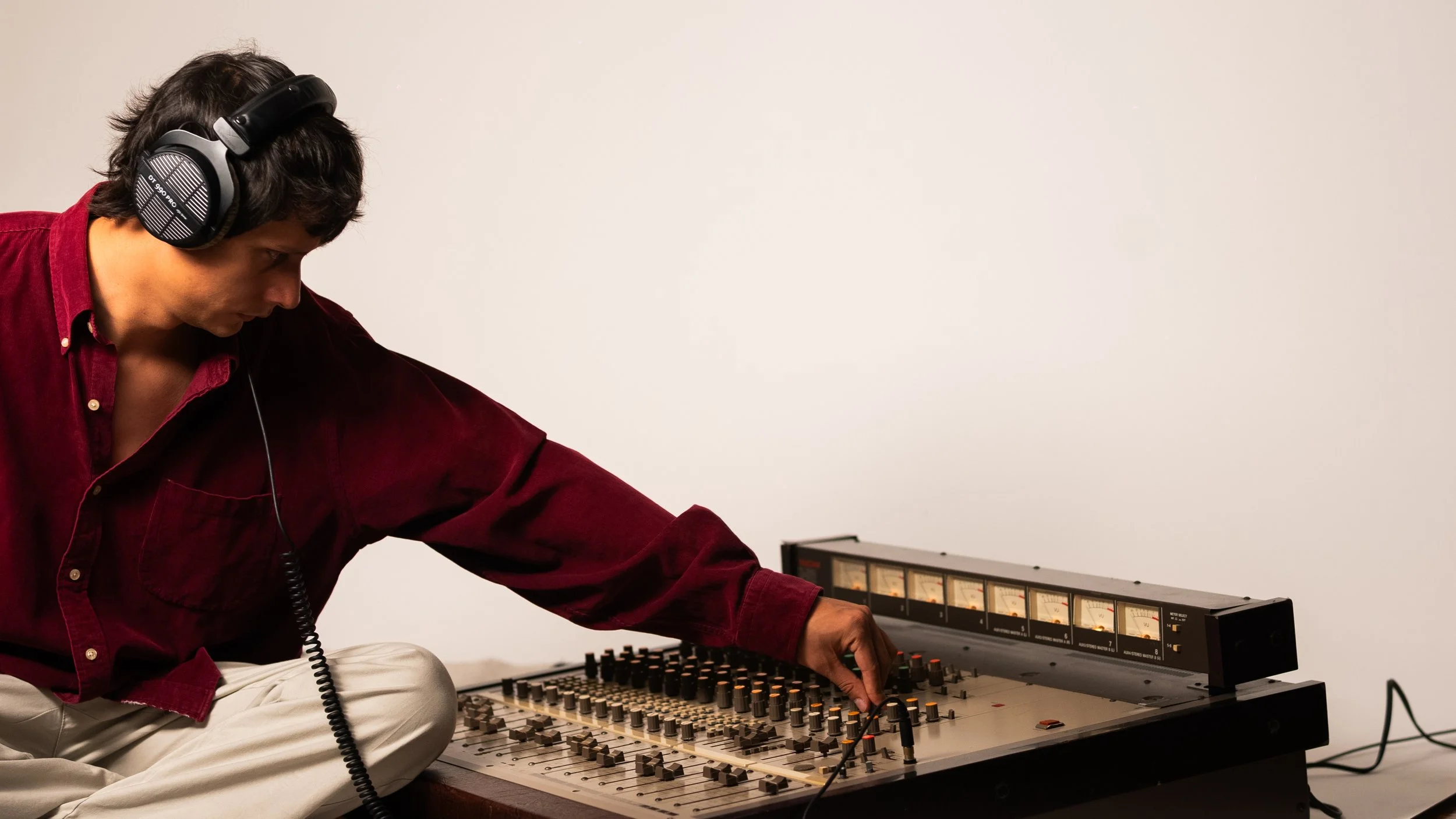 A man wearing a red shirt and beige pants sitting cross-legged and adjusting sliders on a vintage audio mixing console with headphones on.