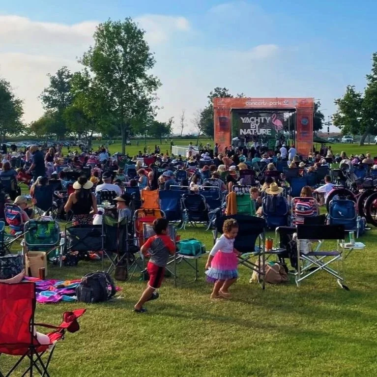 Crowd attending an outdoor concert at a park with band "Yachty by Nature". People are sitting on lawn chairs and blankets, children playing and families dancing and singing.