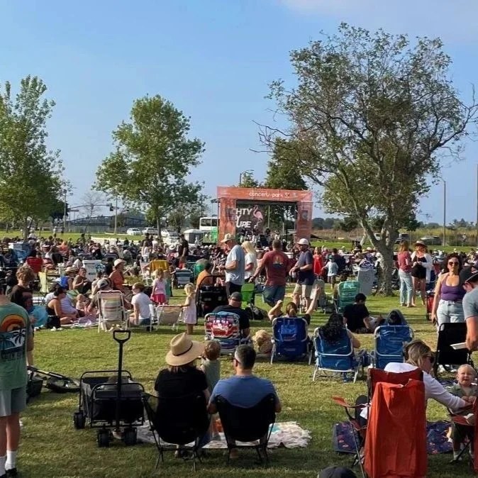 Crowd of people enjoying an outdoor concert in a park, with a stage featuring a band and several large trees providing shade.