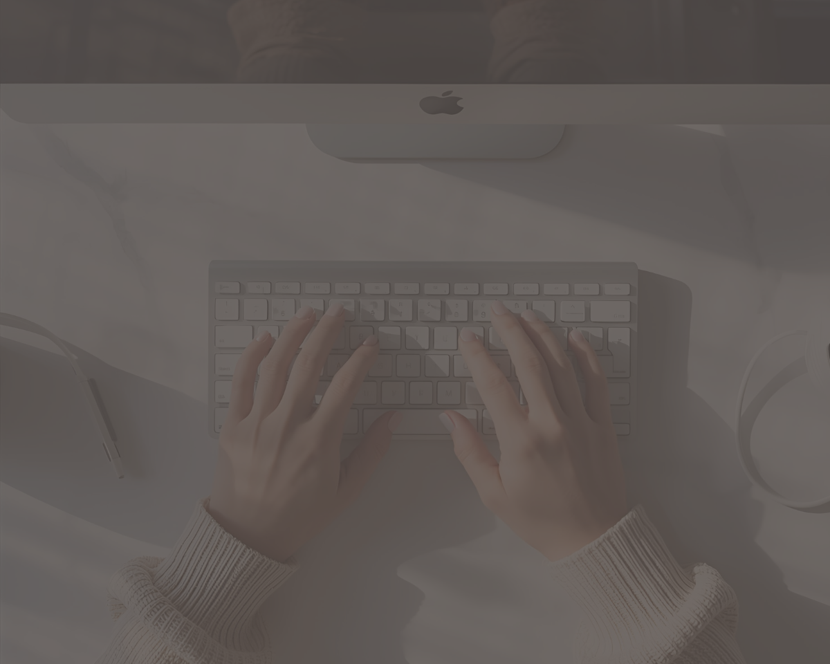Person typing on a white computer keyboard, viewed from above, with a desktop monitor and headphones on a white desk.