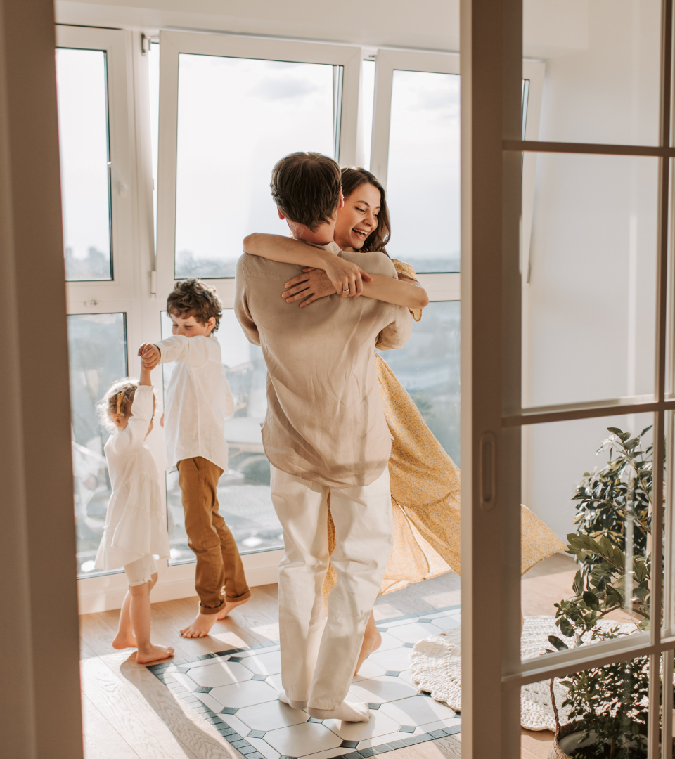 A family dancing and hugging by large windows in a bright living room with three children and an adult woman, with a potted plant visible on the side.
