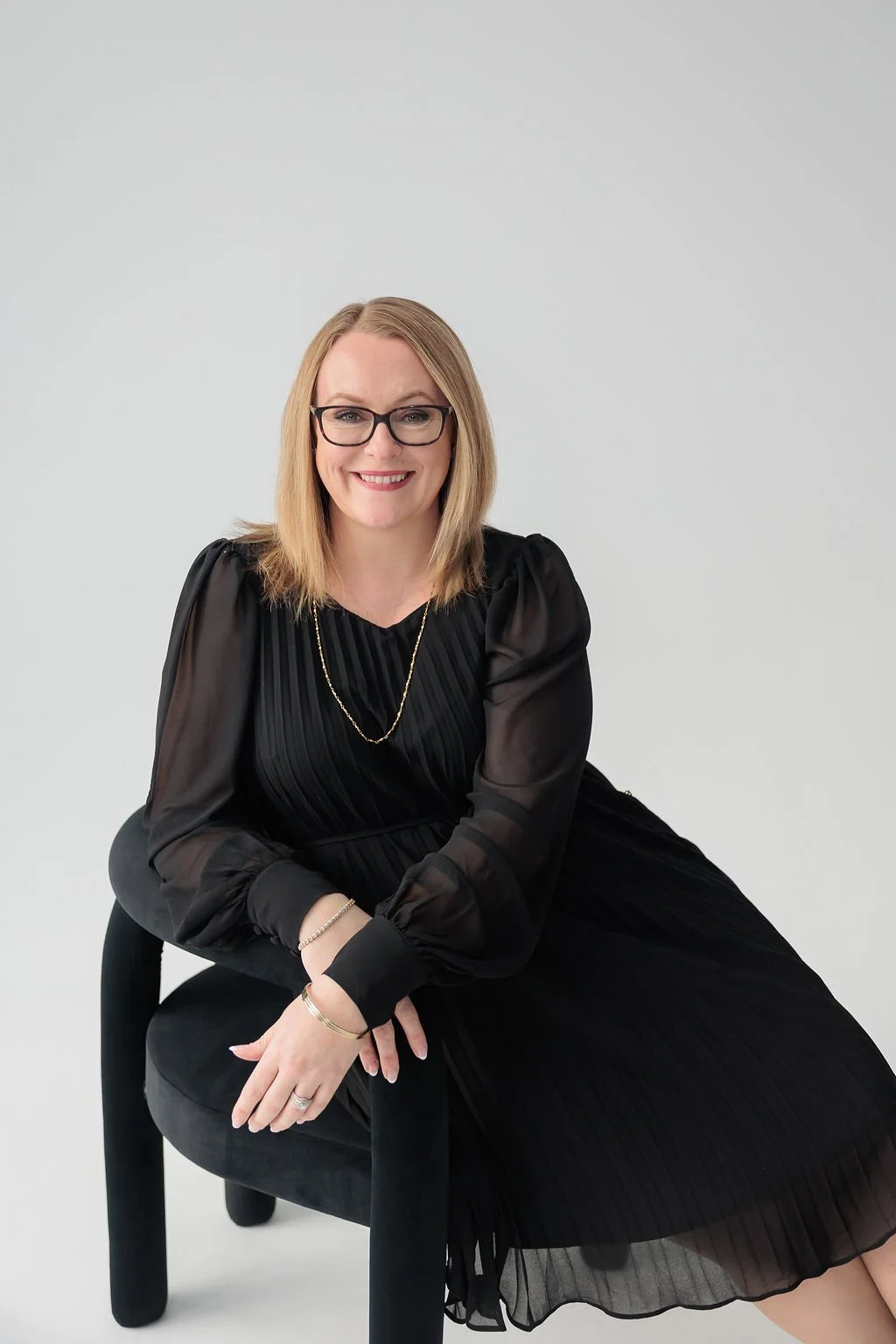 A woman with shoulder-length blonde hair, wearing glasses, a black pleated dress, and jewelry, sitting on a black chair against a plain white background, smiling at the camera.
