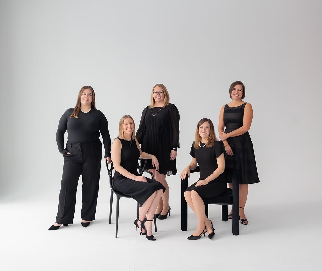 Five women dressed in black, smiling, posing in a white studio background, with some seated on chairs and others standing.