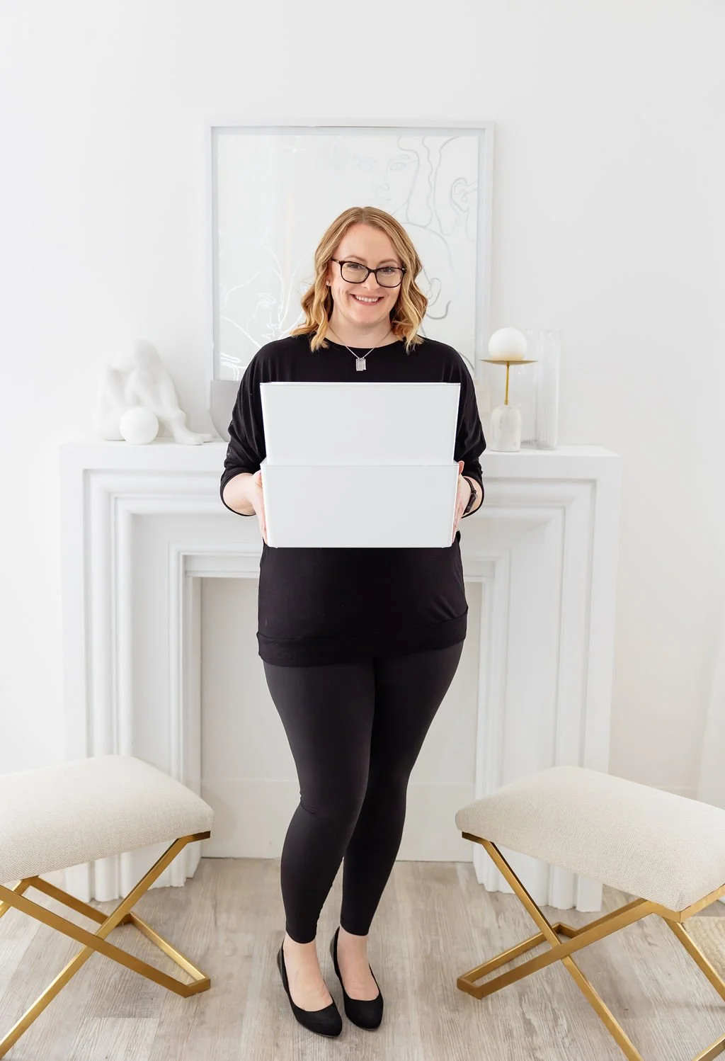 Woman with glasses and blonde hair smiling, holding an open laptop in a bright, modern room with white decor and two beige stools.