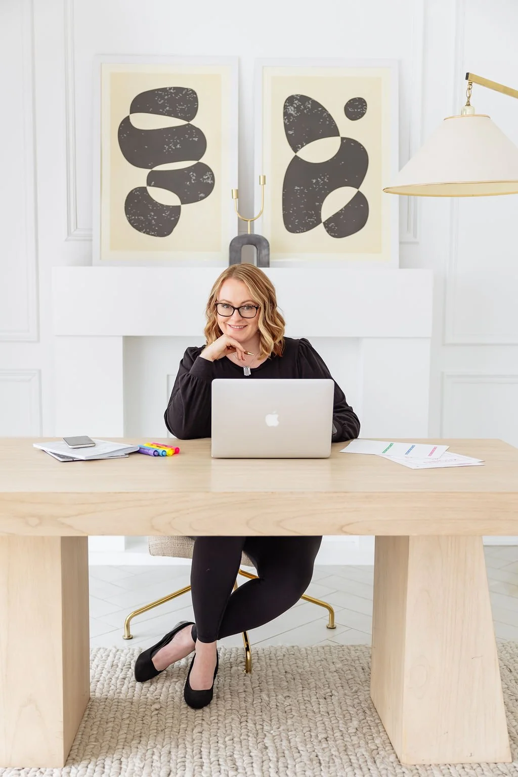 A woman with blonde hair, glasses, and a black top sitting at a light wood desk in a bright, modern office. She has a laptop in front of her, with papers and colorful markers on the desk. Behind her is white wall art with abstract black shapes and a gold candelabra, and a large white-shaded lamp hangs from the ceiling.