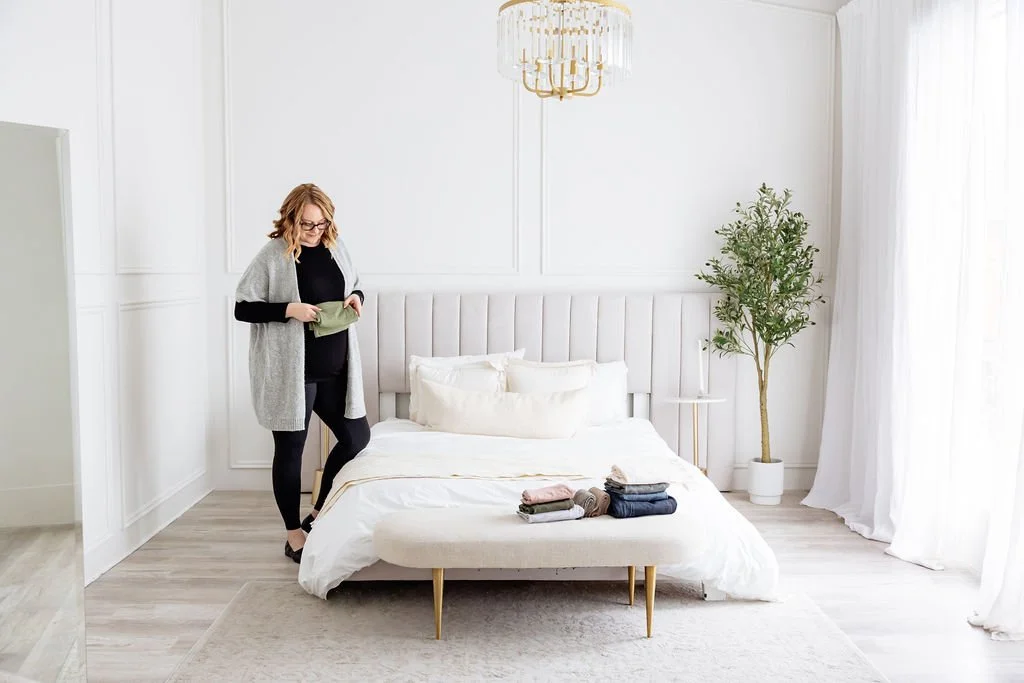 A woman preparing a bed in a bright, minimalist bedroom with white walls, gold chandelier, indoor plant, and floor-to-ceiling curtains.