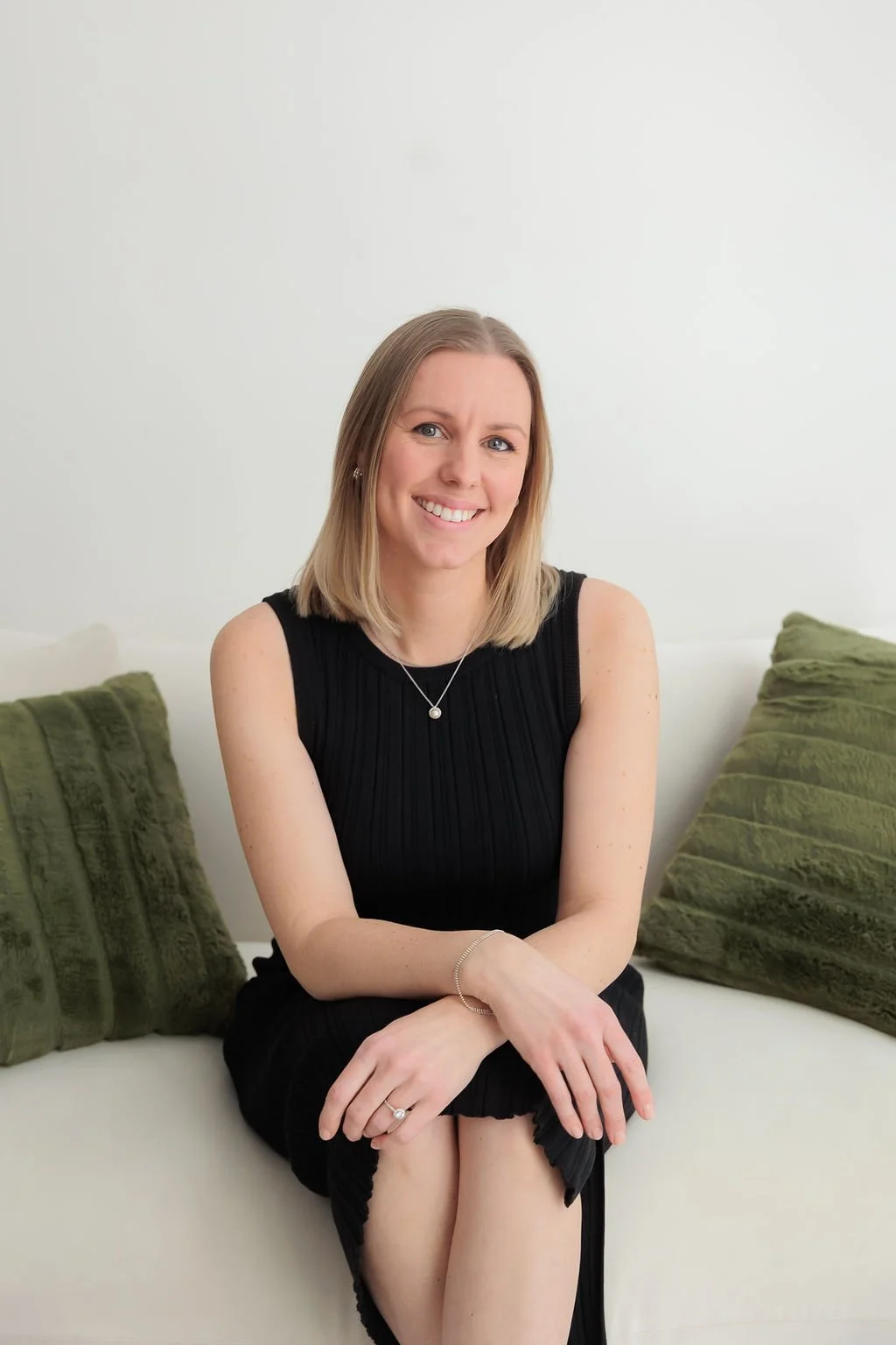 A smiling woman with shoulder-length blonde hair sitting on a white couch with green pillows, wearing a sleeveless black dress and jewelry.