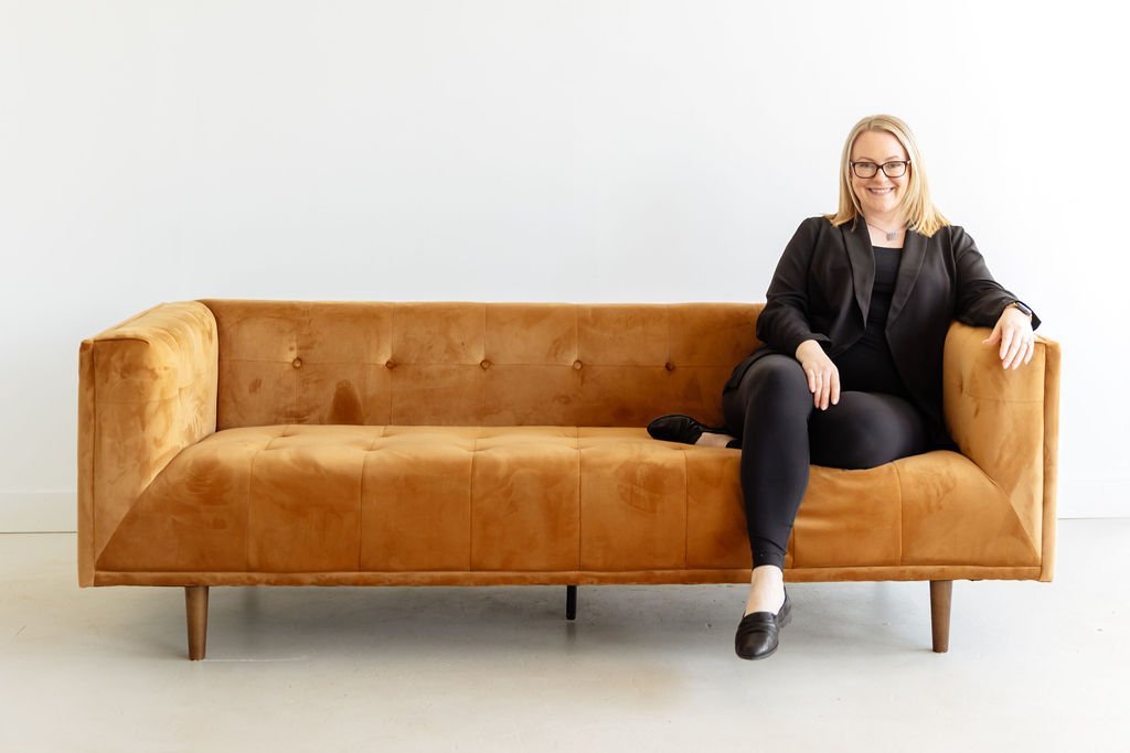 A woman sitting on a tan leather couch against a plain white wall, smiling, dressed in black with glasses.