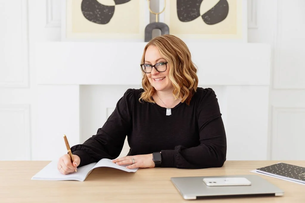 A woman with glasses and wavy red hair sitting at a wooden desk, smiling, holding a pen over an open notebook, with a smartphone and closed planner on the desk.
