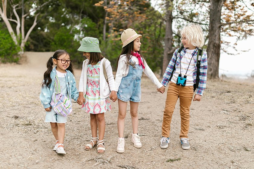 Four young children, two girls and two boys, walking outdoors in a park holding hands and smiling.