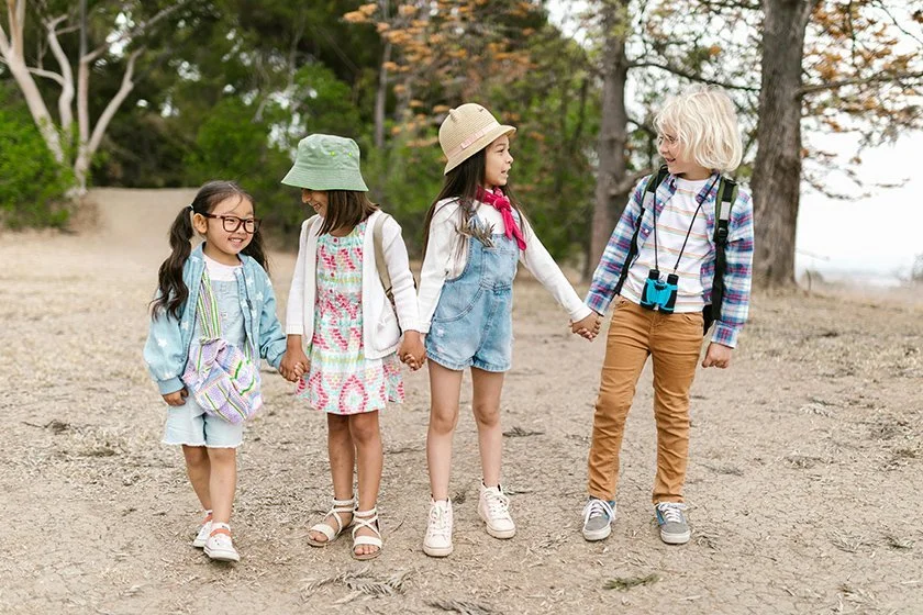 Four children hold hands and walk outdoors in a park with trees in the background.