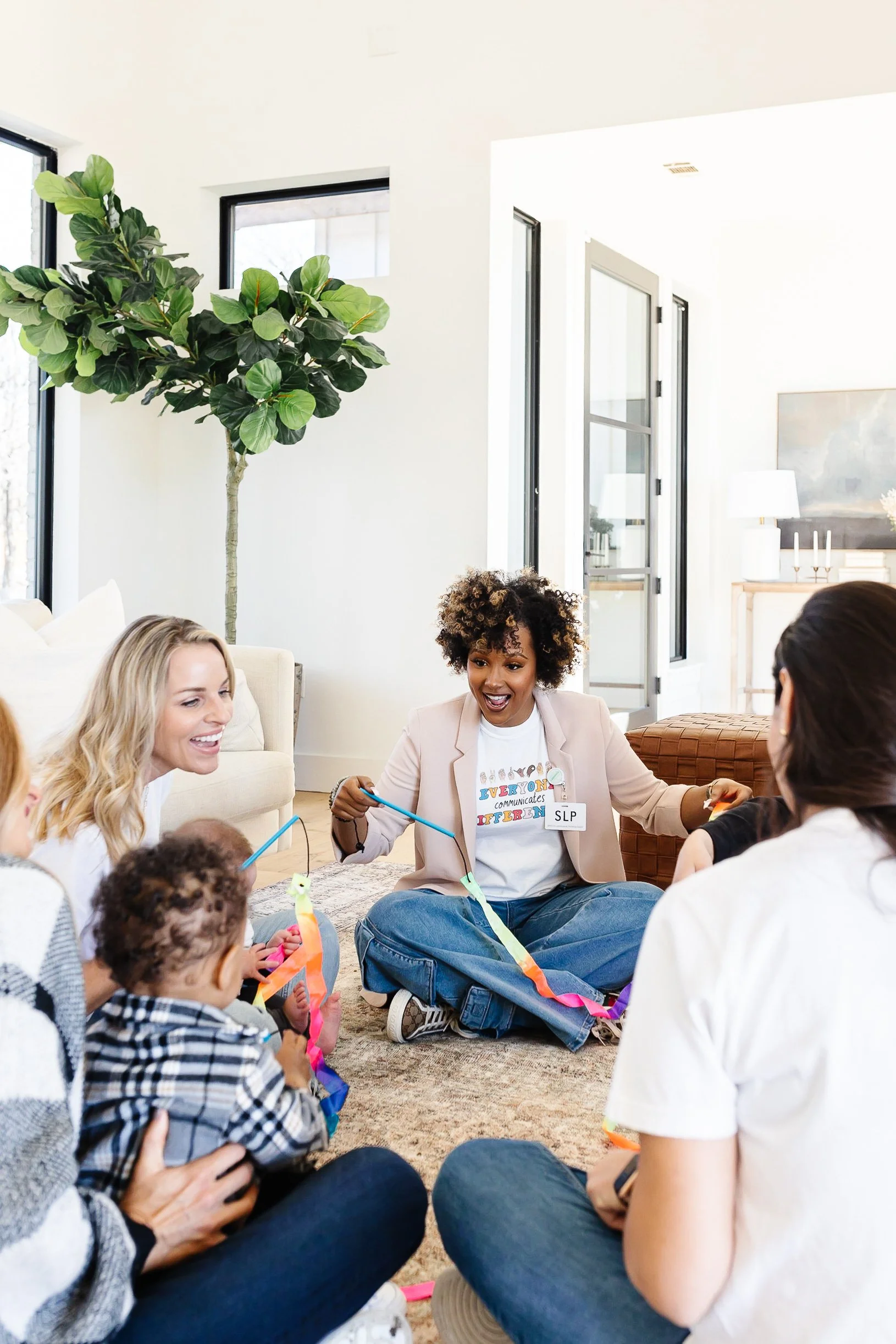 Group of women and children sitting on the floor in a living room, engaging in a colorful craft activity, with the woman in the center leading the activity.