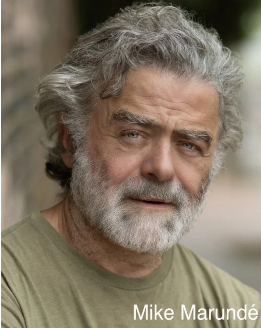 Close-up of an elderly man with gray hair and beard, wearing a green shirt, outdoors, with the name 'Mike Marundé' captioned at the bottom.