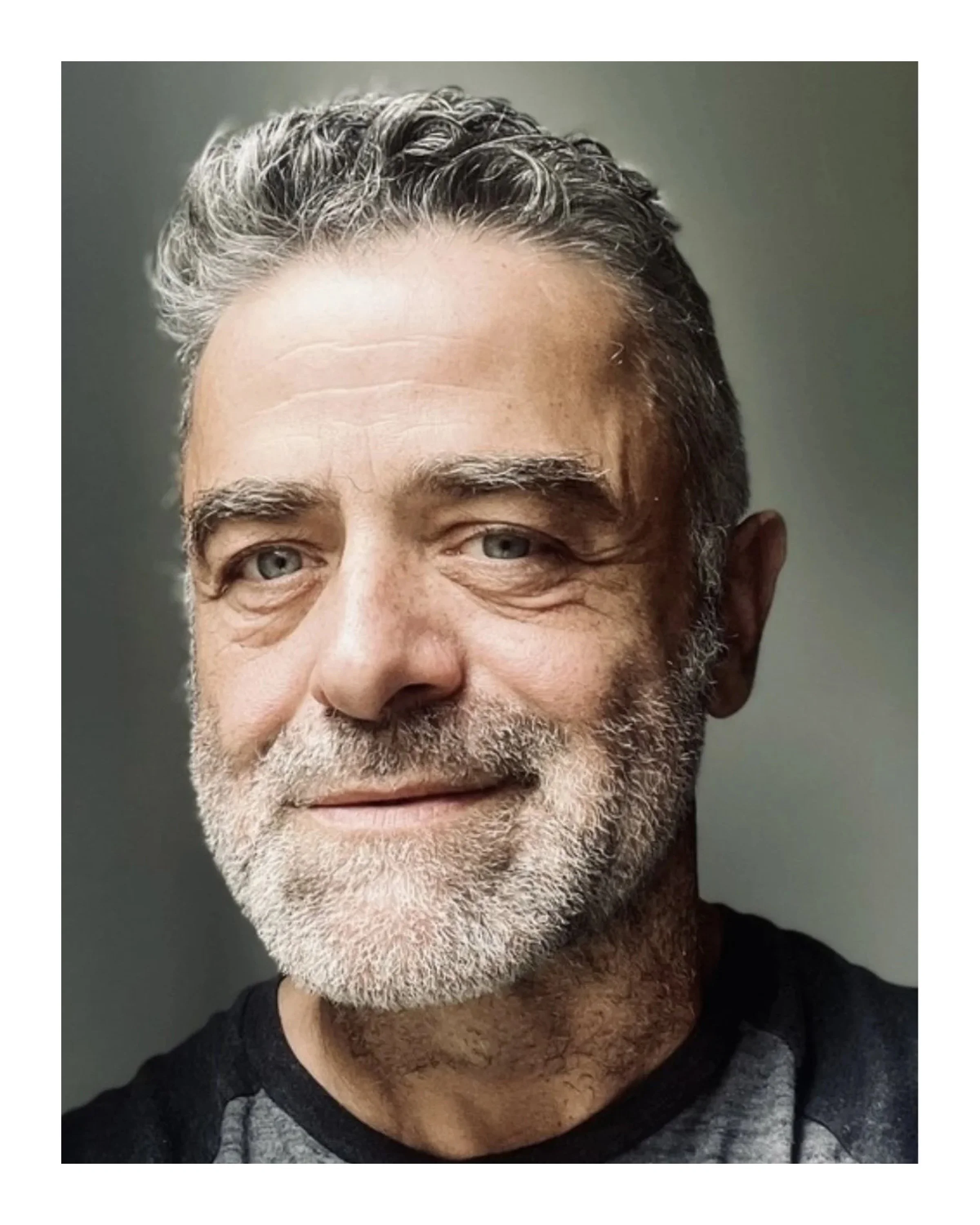 Close-up of an older man's face with gray hair, gray beard, and blue eyes, smiling slightly, wearing a black and gray shirt, against a plain background.
