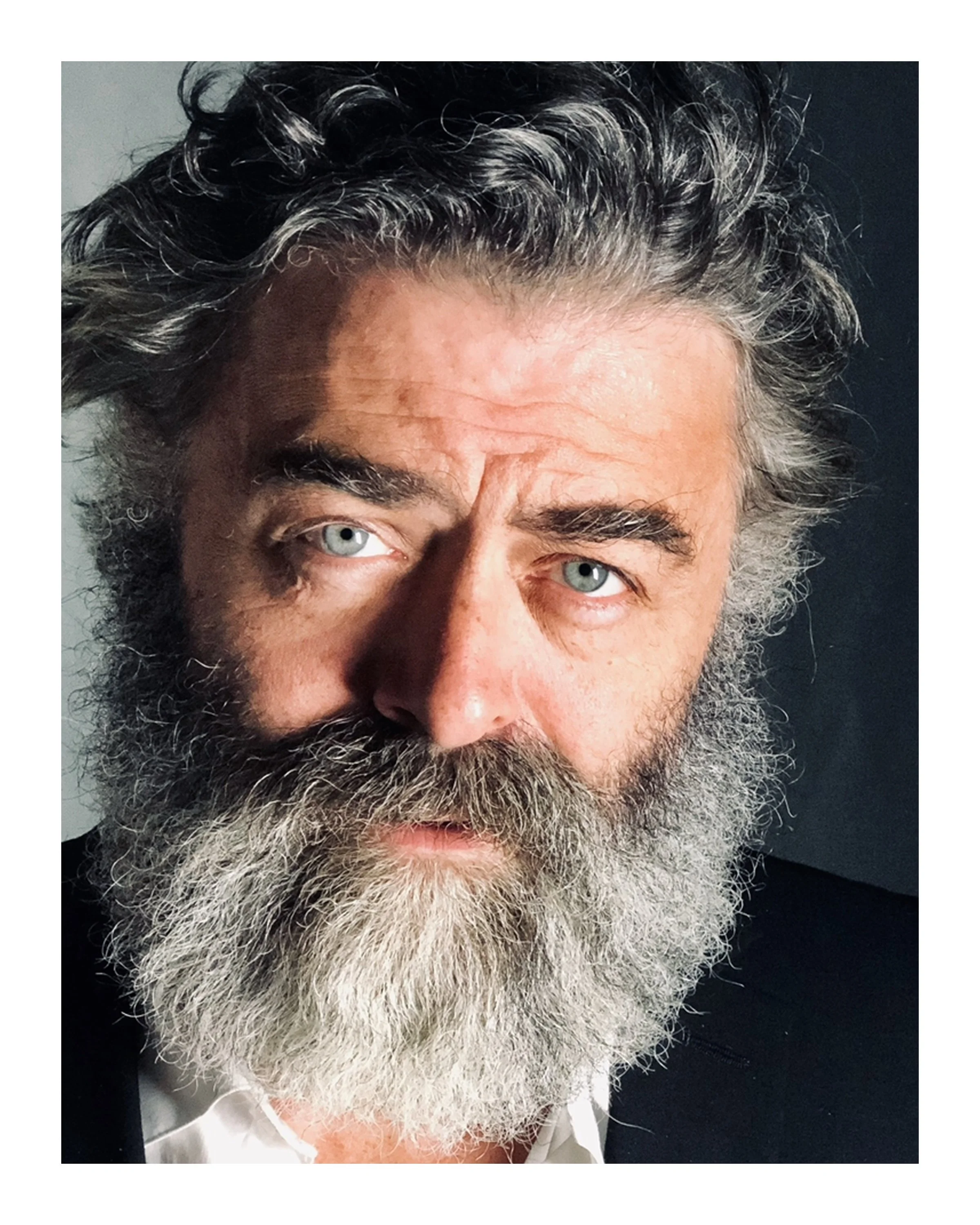 Close-up of a man with gray hair and beard, light-colored eyes, wearing a white shirt and dark jacket, against a contrasting dark background.