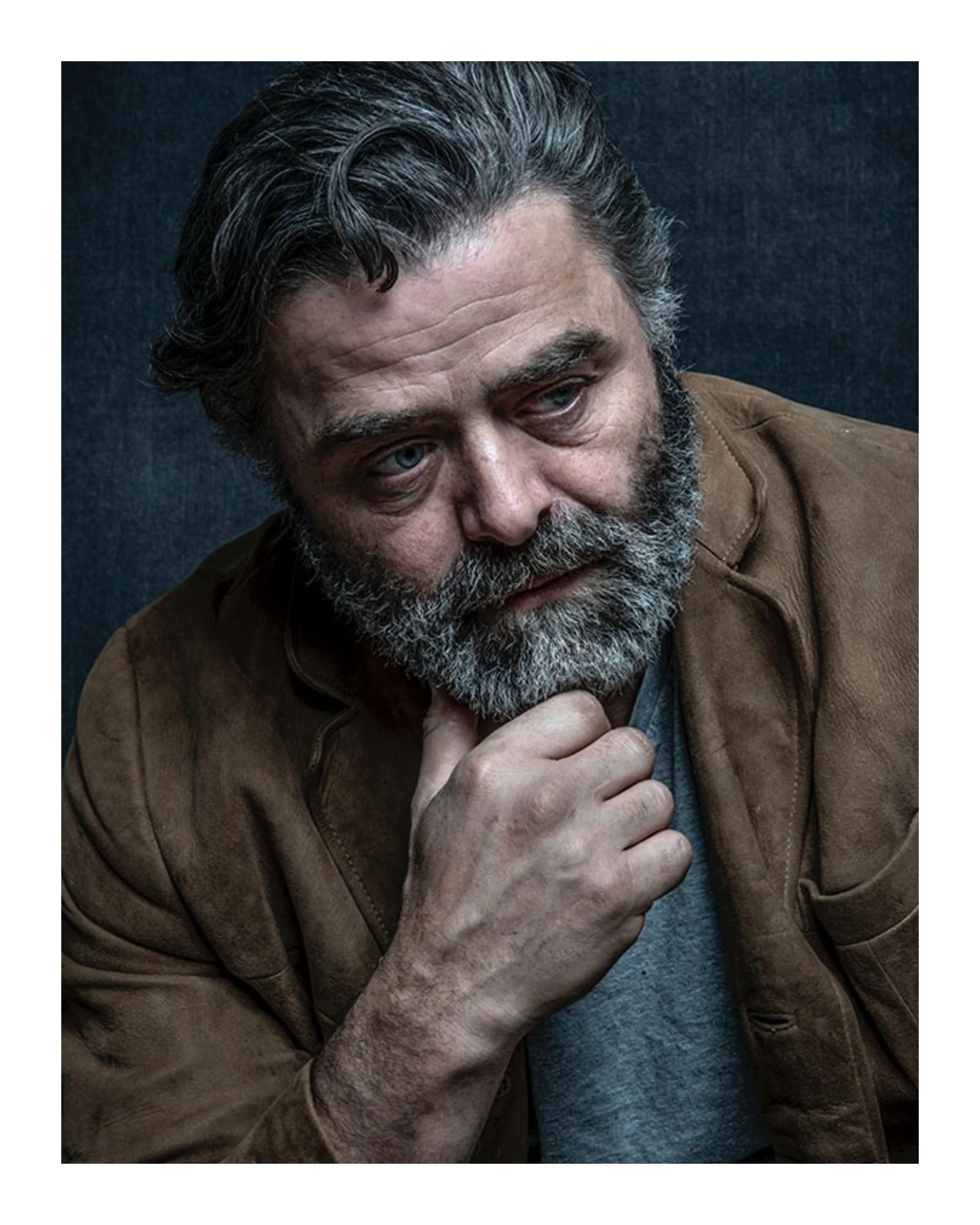 A man with gray hair and a beard, wearing a brown jacket, rests his chin on his hand and looks thoughtful against a dark background.