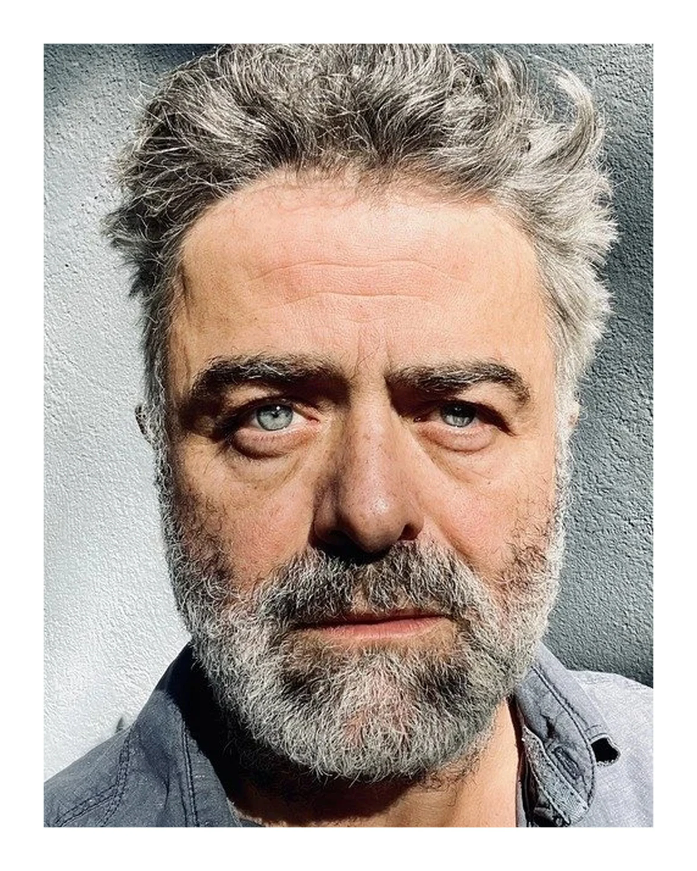 Close-up of a middle-aged man with gray hair, blue eyes, and a gray beard, wearing a gray shirt, standing against a textured wall.