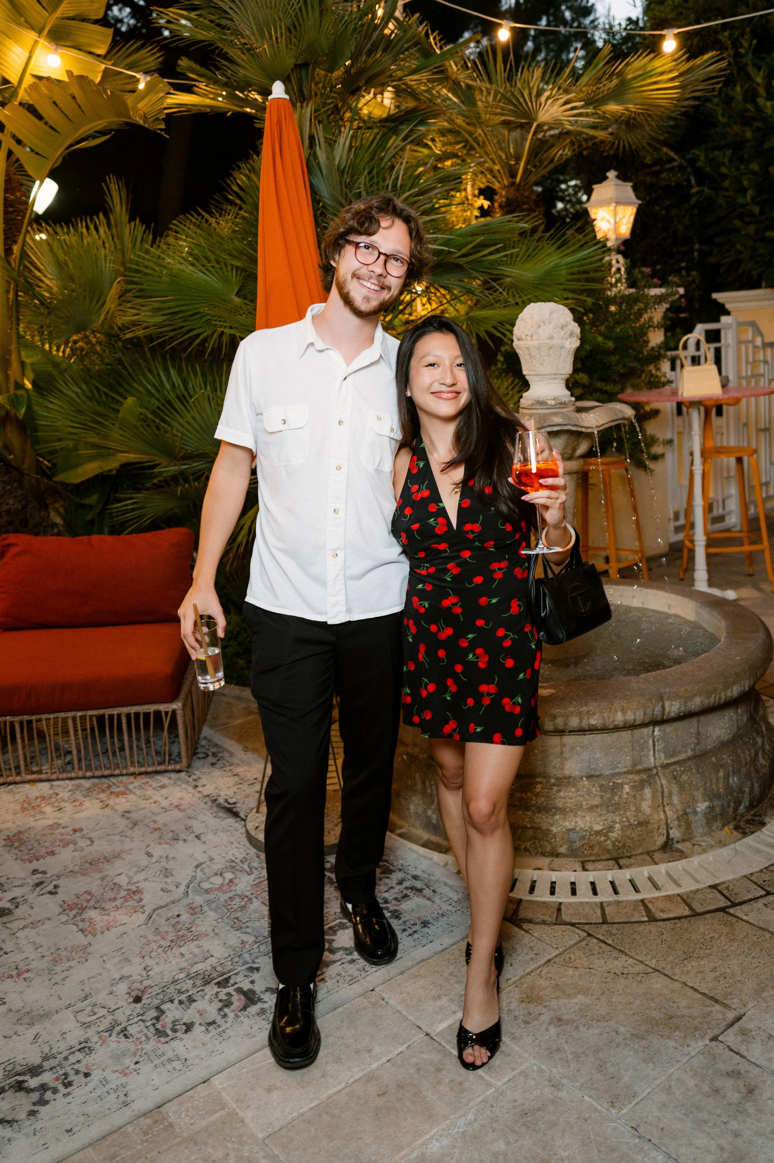 A man and woman smiling and standing together outdoors in the evening. The woman is holding a glass of rosé wine, and the man is holding a glass of clear beverage. They are standing near a fountain with lush tropical plants and string lights overhead.