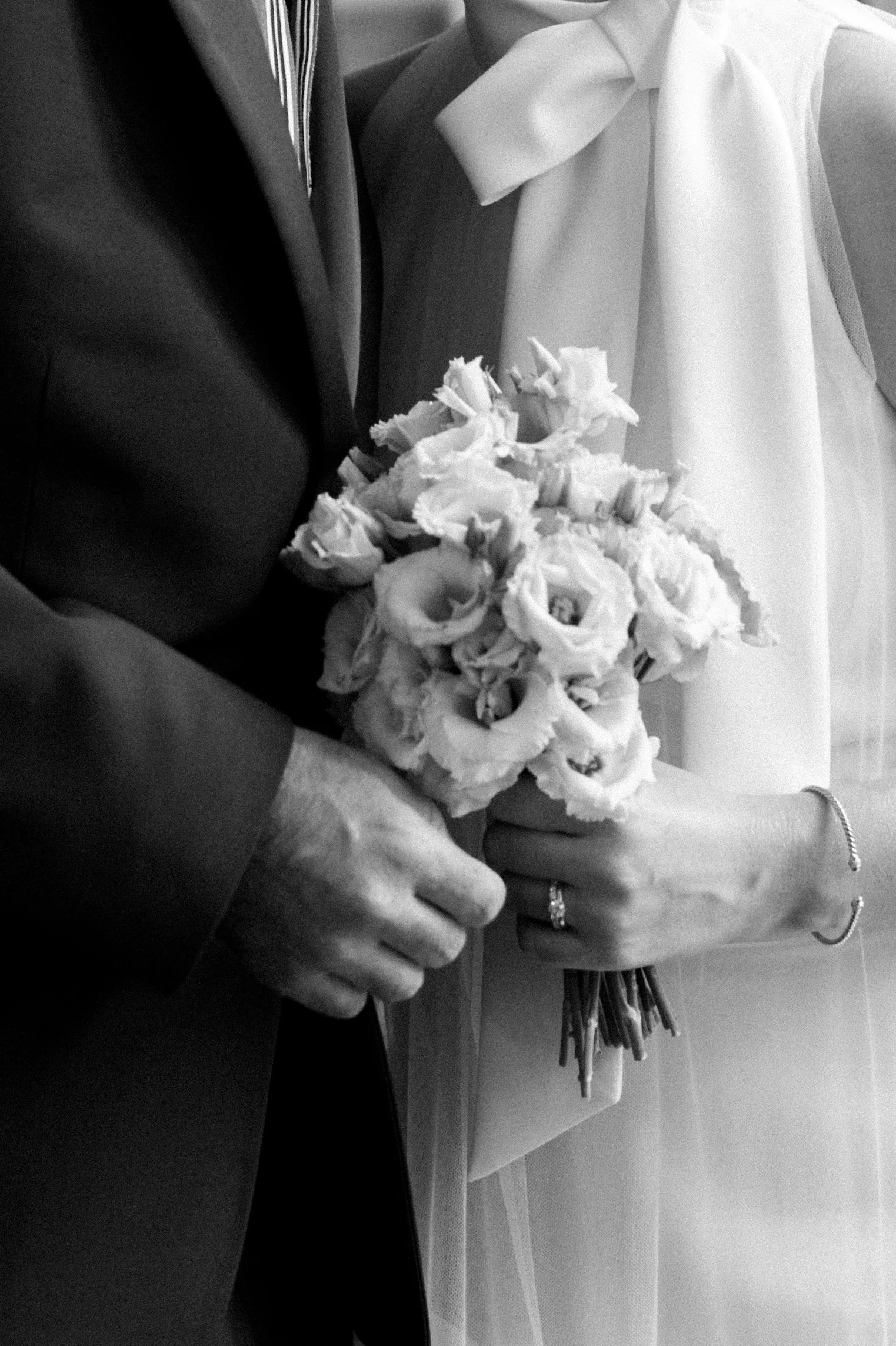Close-up of a bride and groom holding a flower bouquet, with the groom's hand and the bride's hand visible, showing wedding rings.