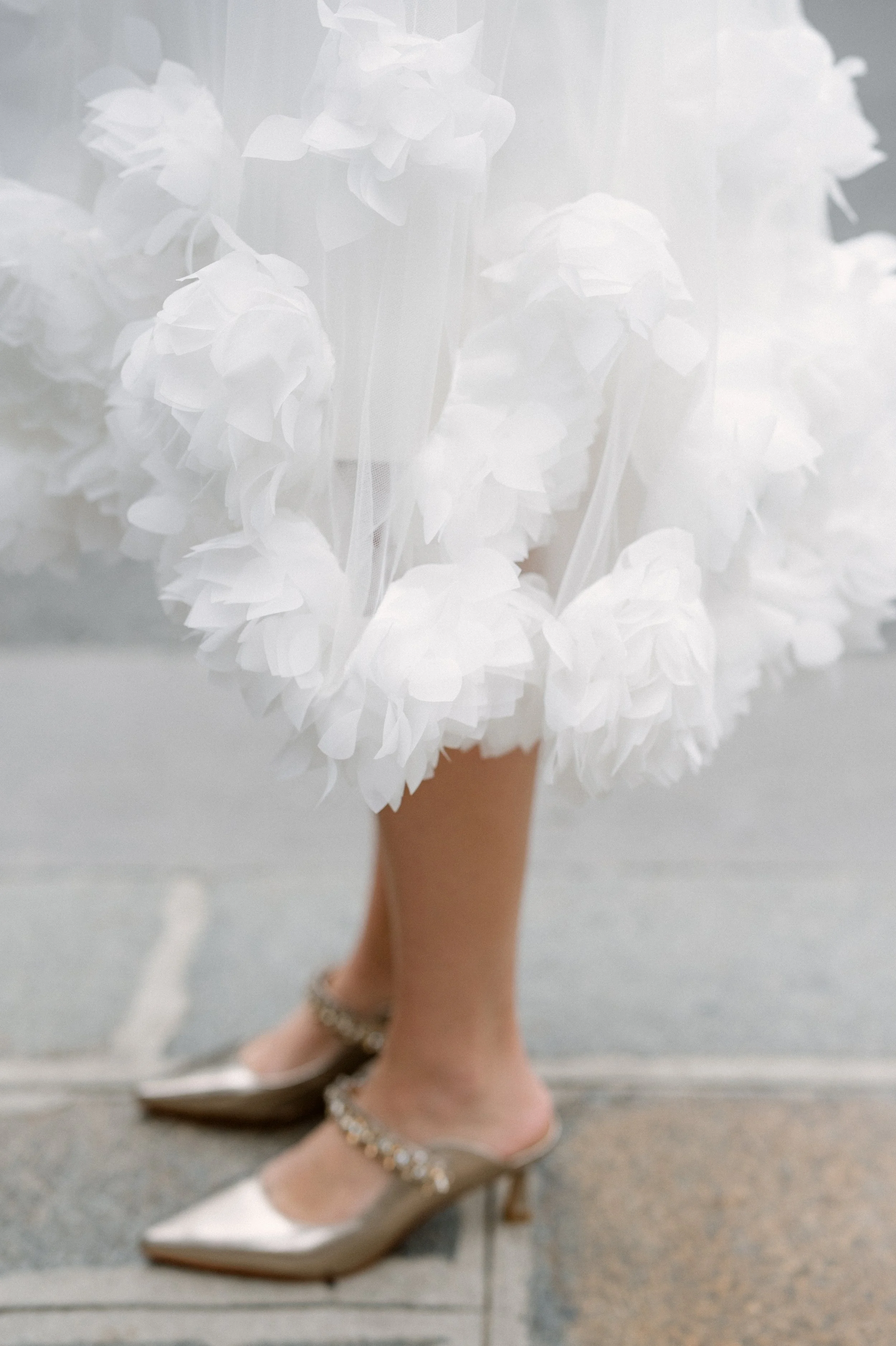 Close-up of a person's legs and feet in high-heeled shoes, with a white, frilly dress or skirt covering the upper part of the legs. The shoes are metallic gold with embellishments around the ankle area.