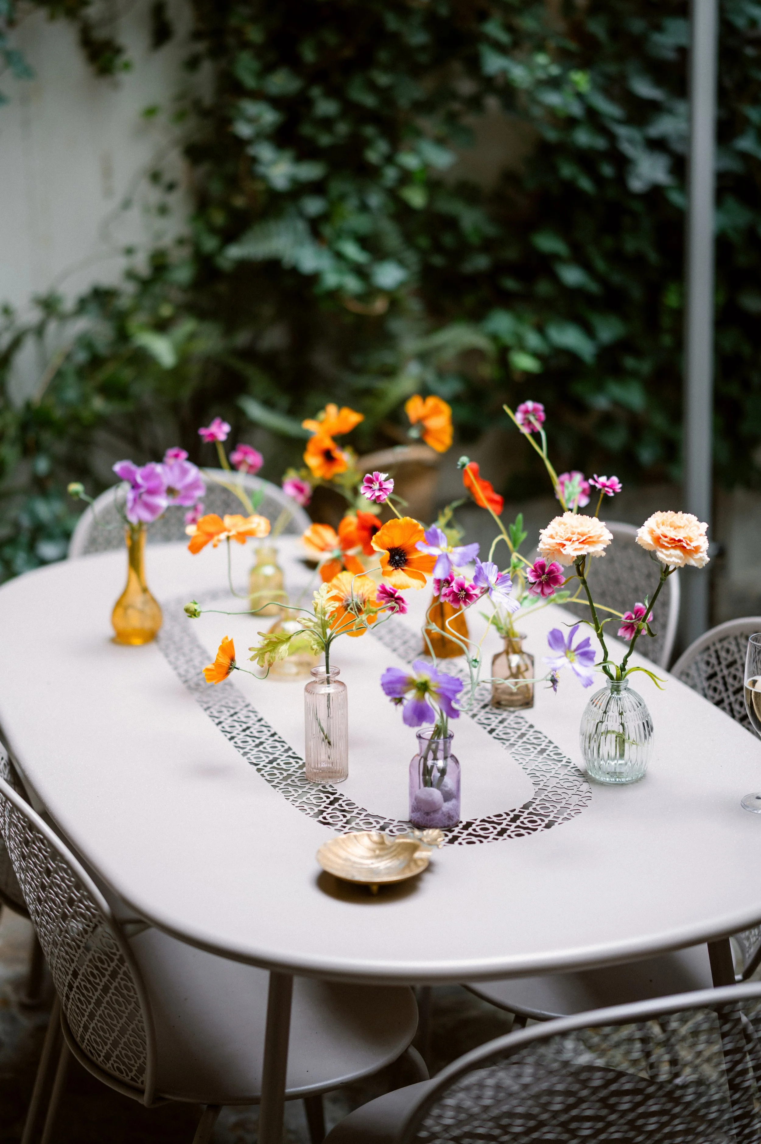 A white outdoor dining table decorated with colorful flower arrangements in glass vases, a lace table runner, and small decorative items, surrounded by white chairs with intricate designs and greenery in the background.
