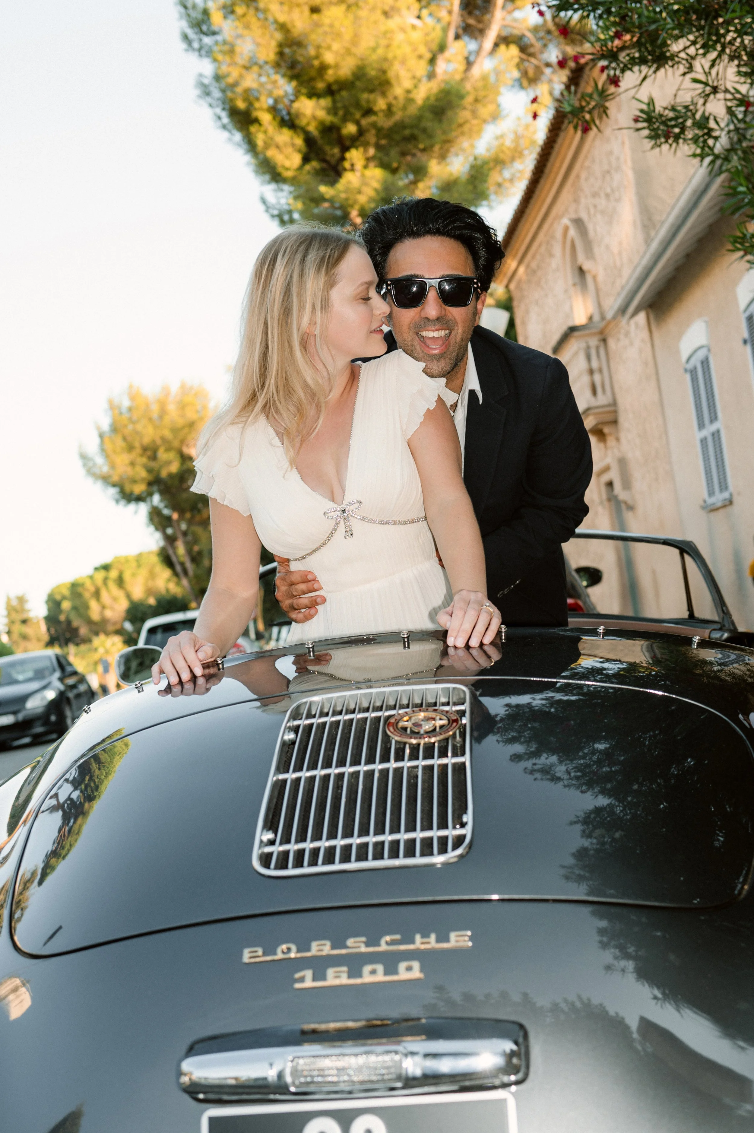 A man and woman sitting on the hood of a black Porsche 1600 convertible, smiling and enjoying a sunny day. The woman is whispering to the man, who is wearing sunglasses. There are trees and houses in the background.
