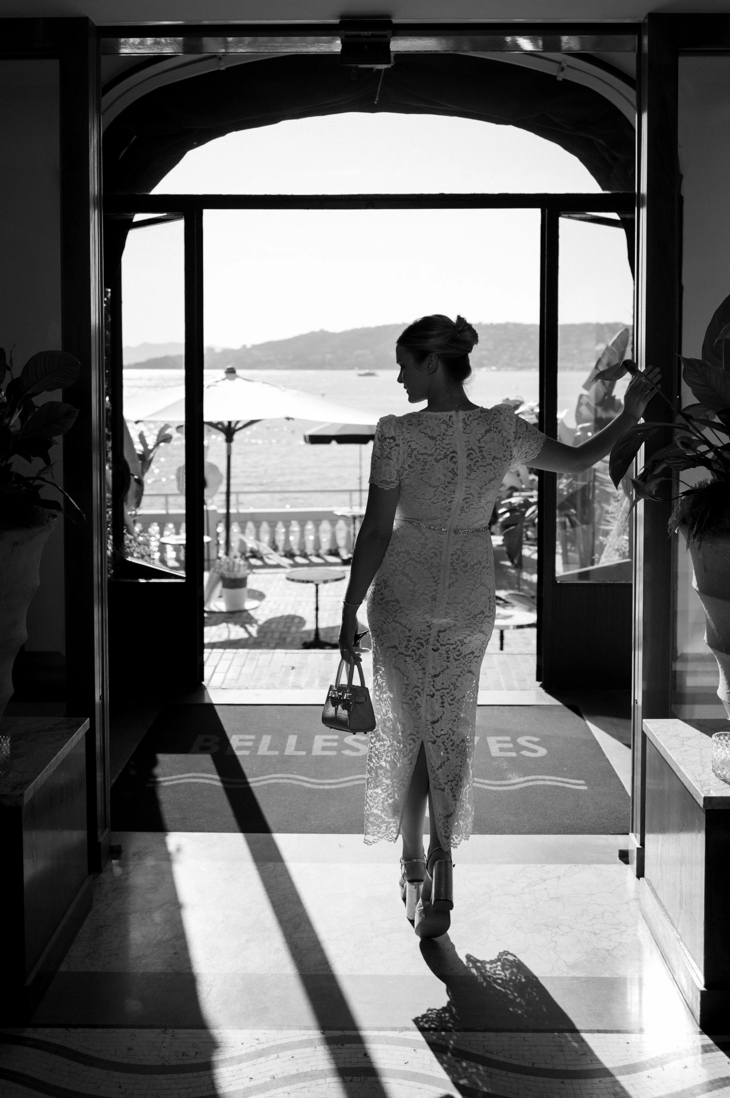 A woman in a lace dress carrying a handbag, walking out of a hotel or restaurant with a view of water and umbrellas outside.