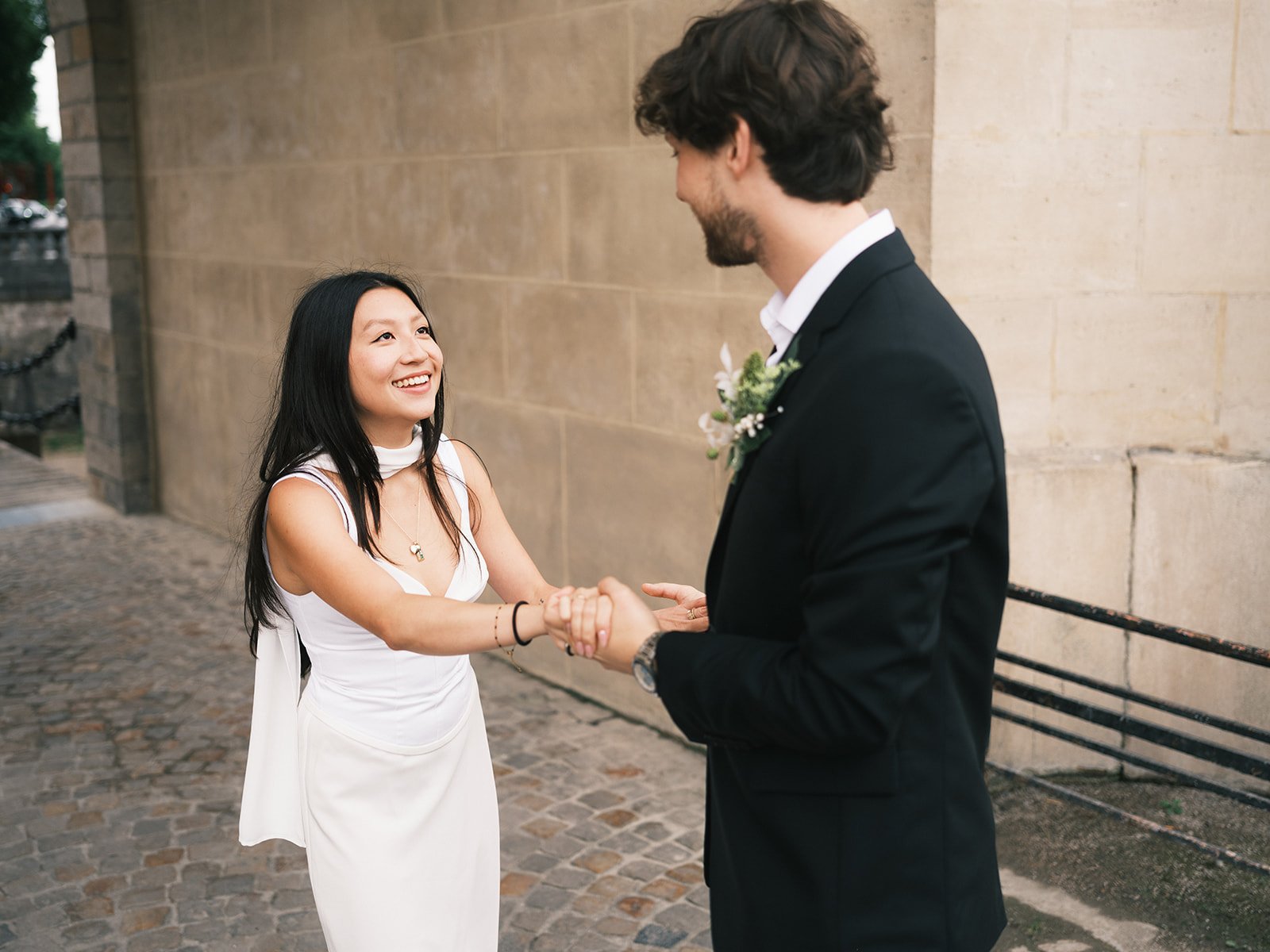 A woman in a white dress smiling and holding hands with a man in a black suit outside next to a stone wall.