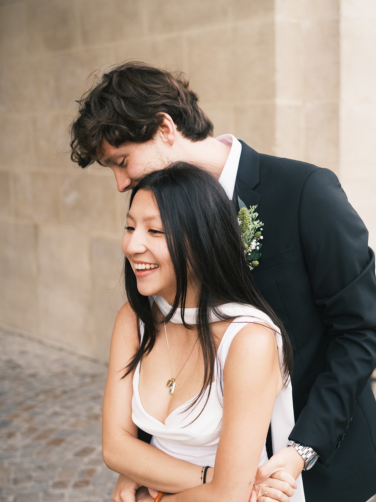 A couple, a man and a woman, sharing a tender moment outdoors. The man, dressed in a black suit with a boutonniere, is leaning over the woman, who is wearing a white dress with a necklace, smiling happily.