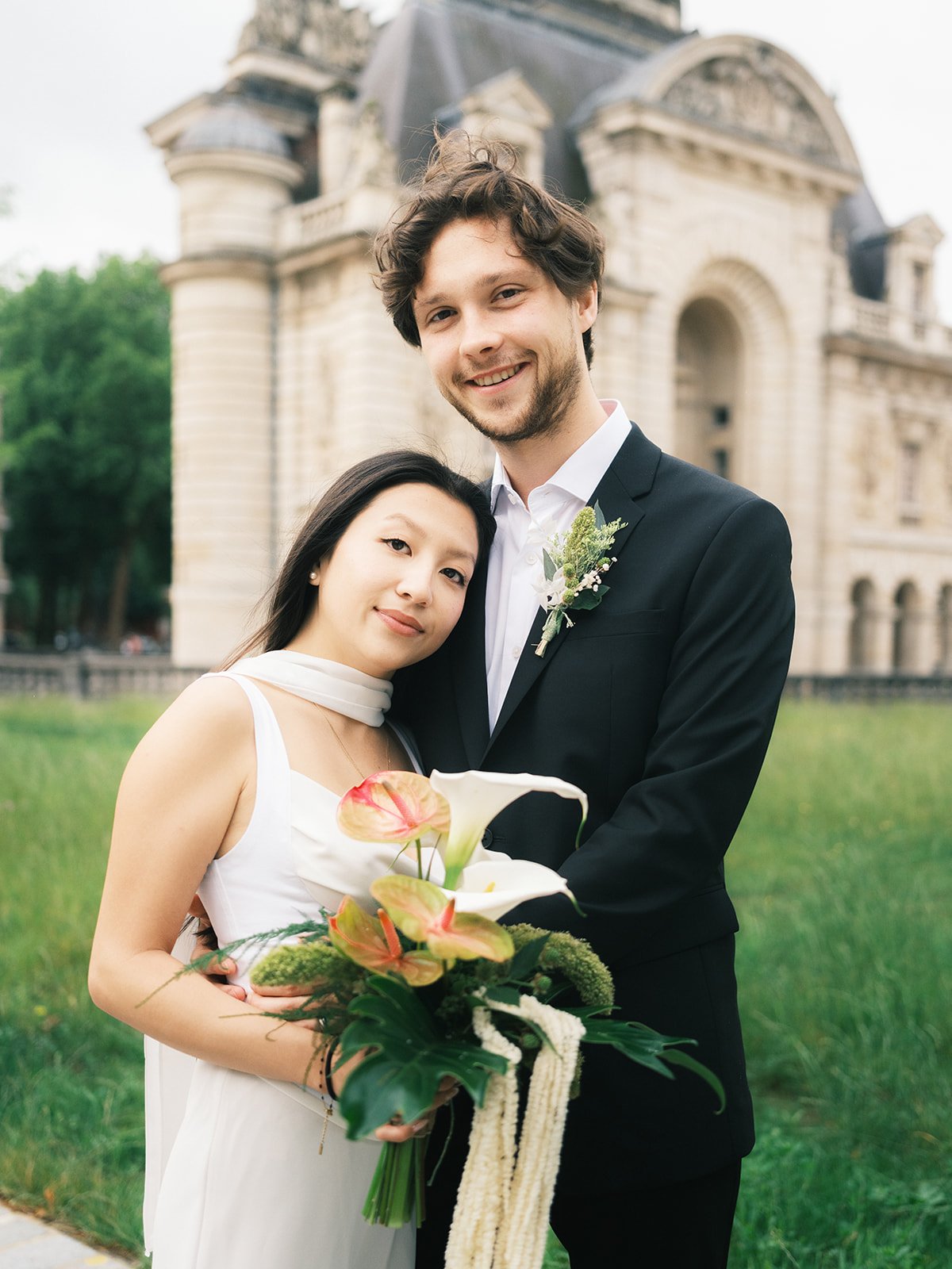A newlywed couple standing outside in front of a historic building, with the bride holding a bouquet of flowers and the groom wearing a boutonniere, both smiling.