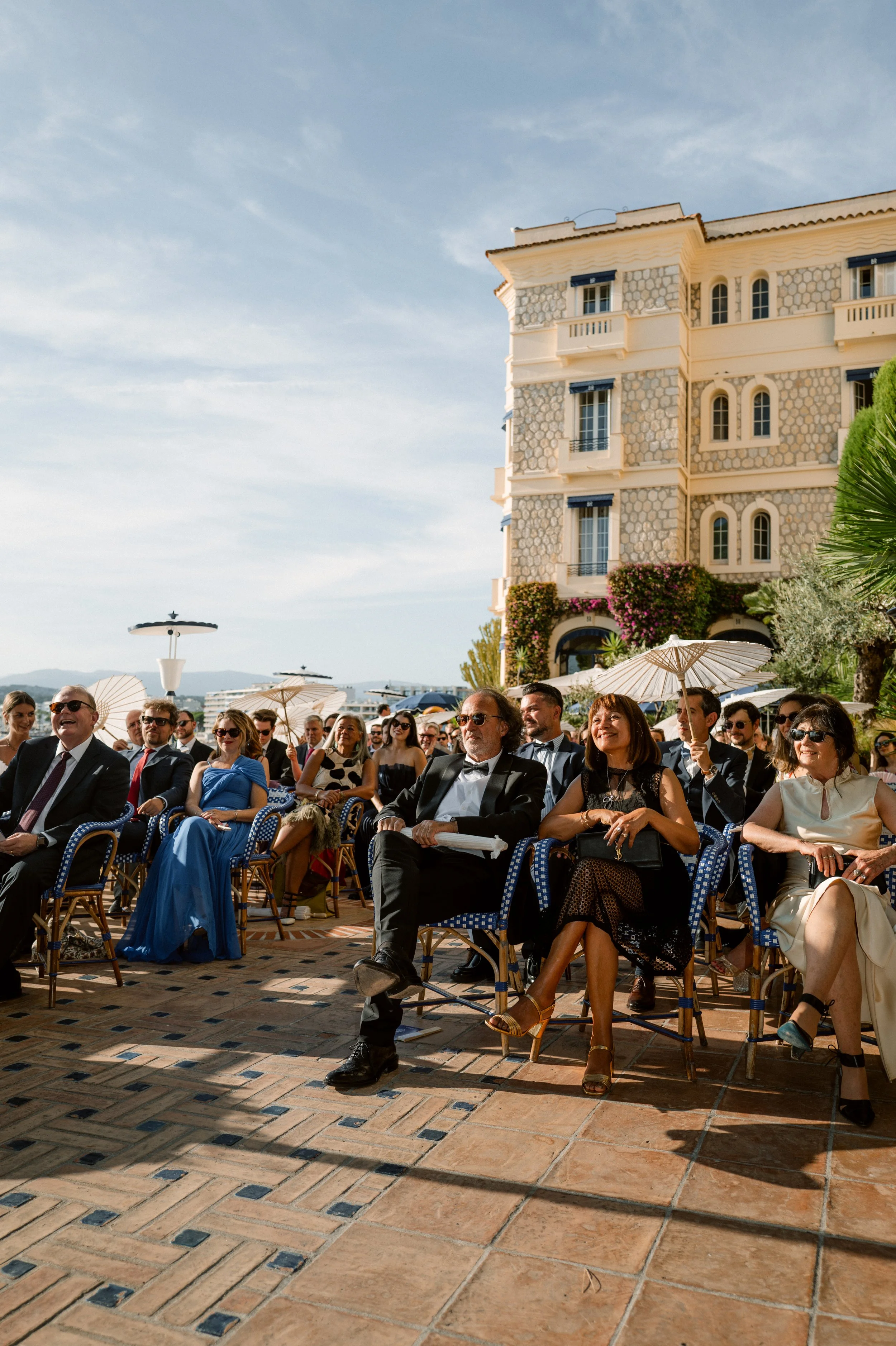 People attending an outdoor wedding ceremony, seated in chairs under umbrellas, in front of a yellow building with balconies and decorative stonework.