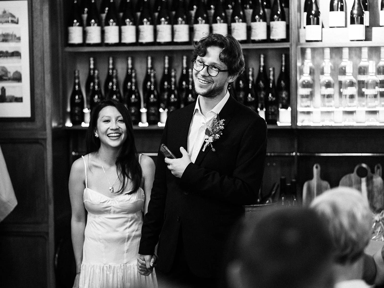 A black and white photo of a smiling couple holding hands at their wedding reception, with bottles of wine on shelves behind them.