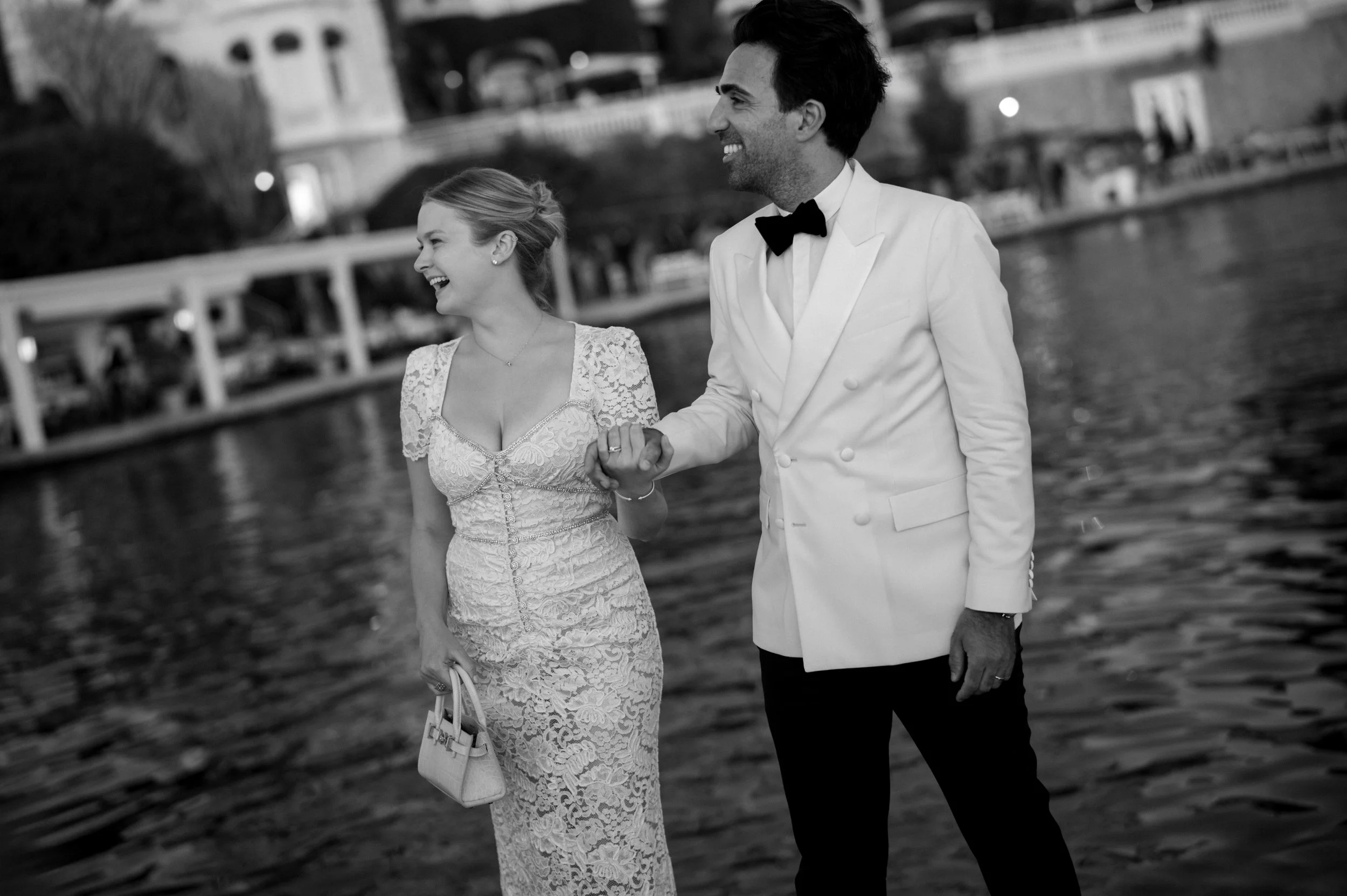 A couple dressed in formal attire, holding hands and smiling, walking near a body of water