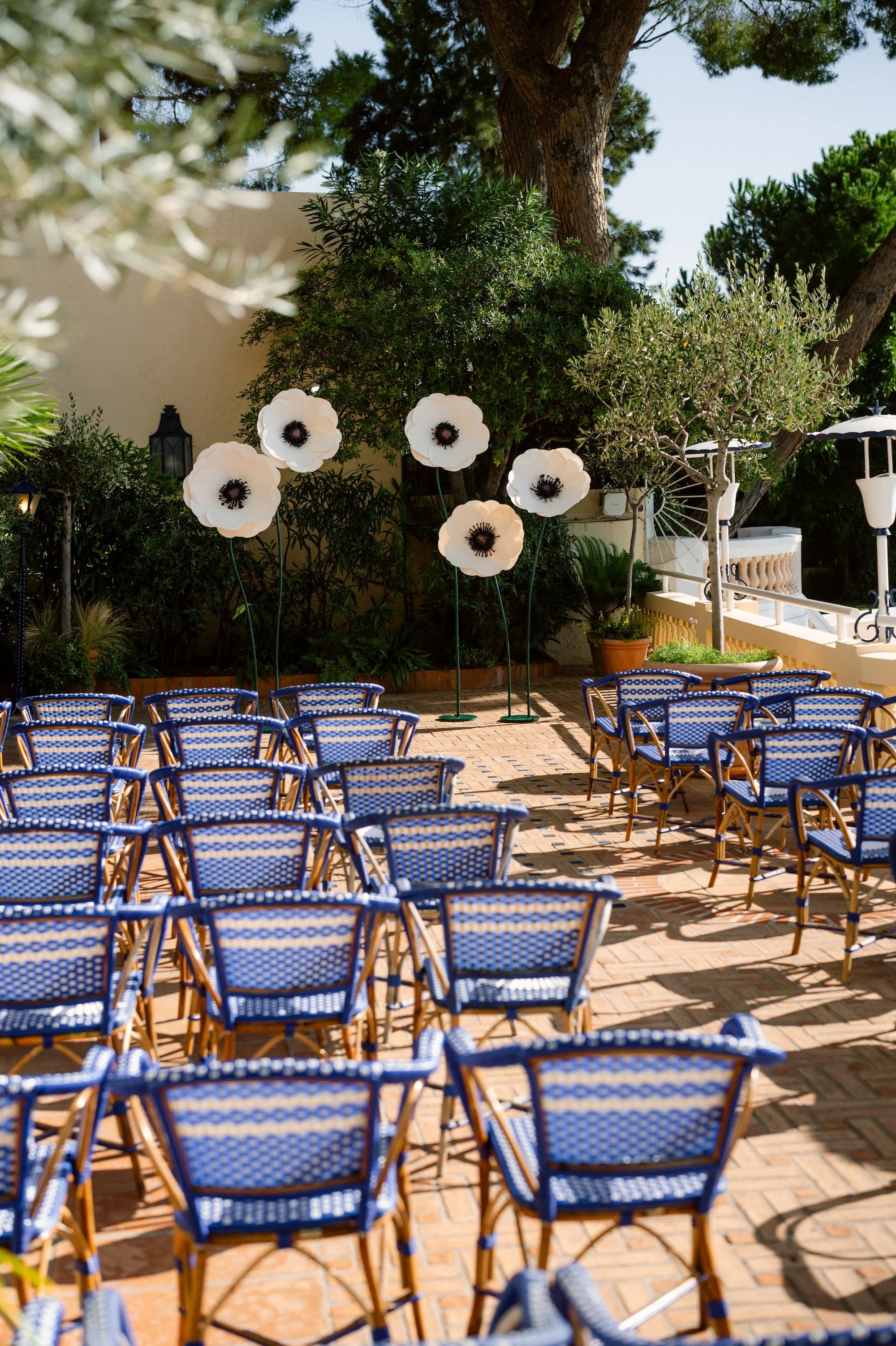 Outdoor patio with rows of blue bistro chairs, large artificial white flowers on tall stems, green trees and bushes, and a staircase with white railings in the background.