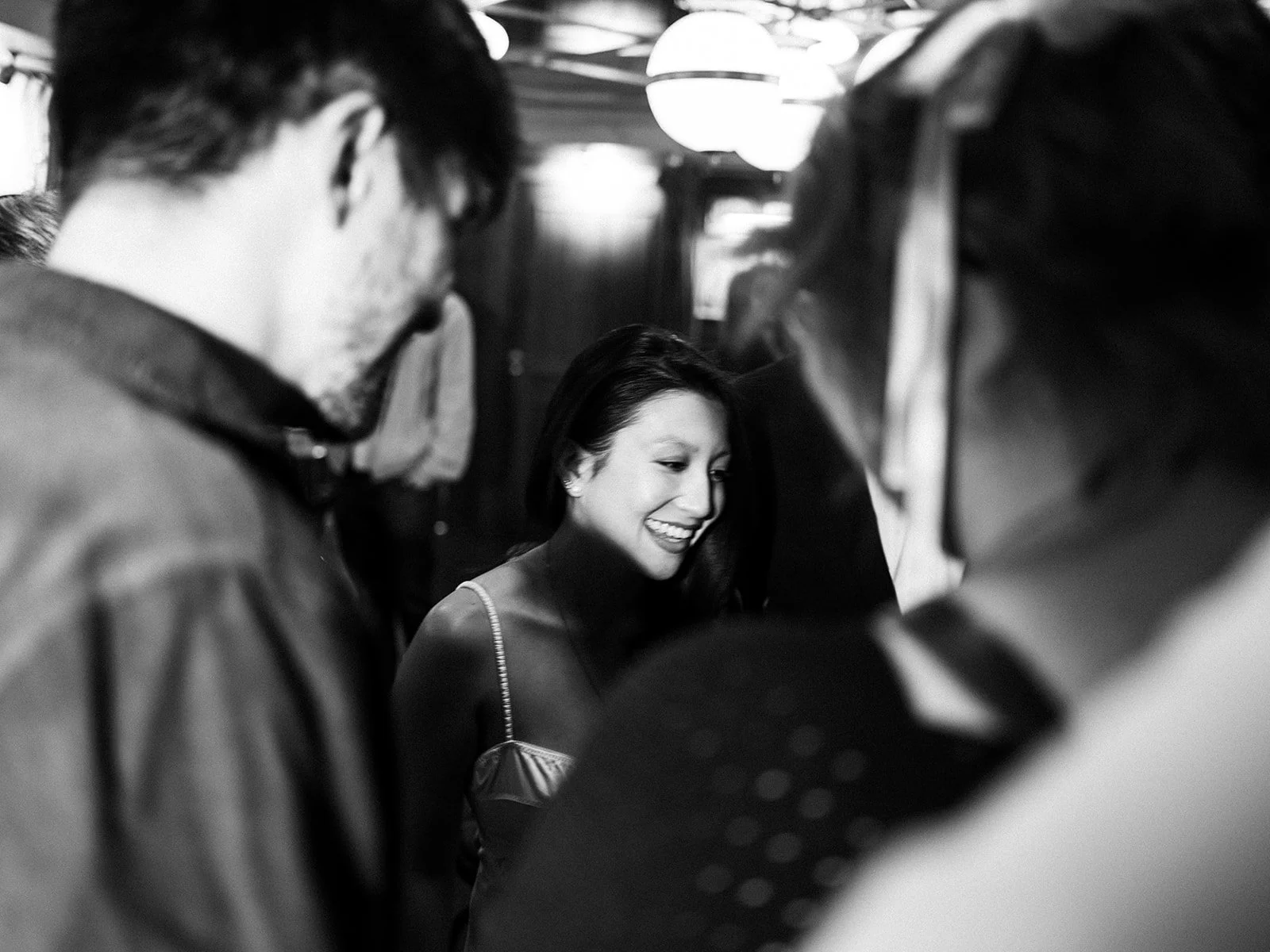 A woman smiling and talking to two men in a social gathering or event, with warm lighting and people in the background.