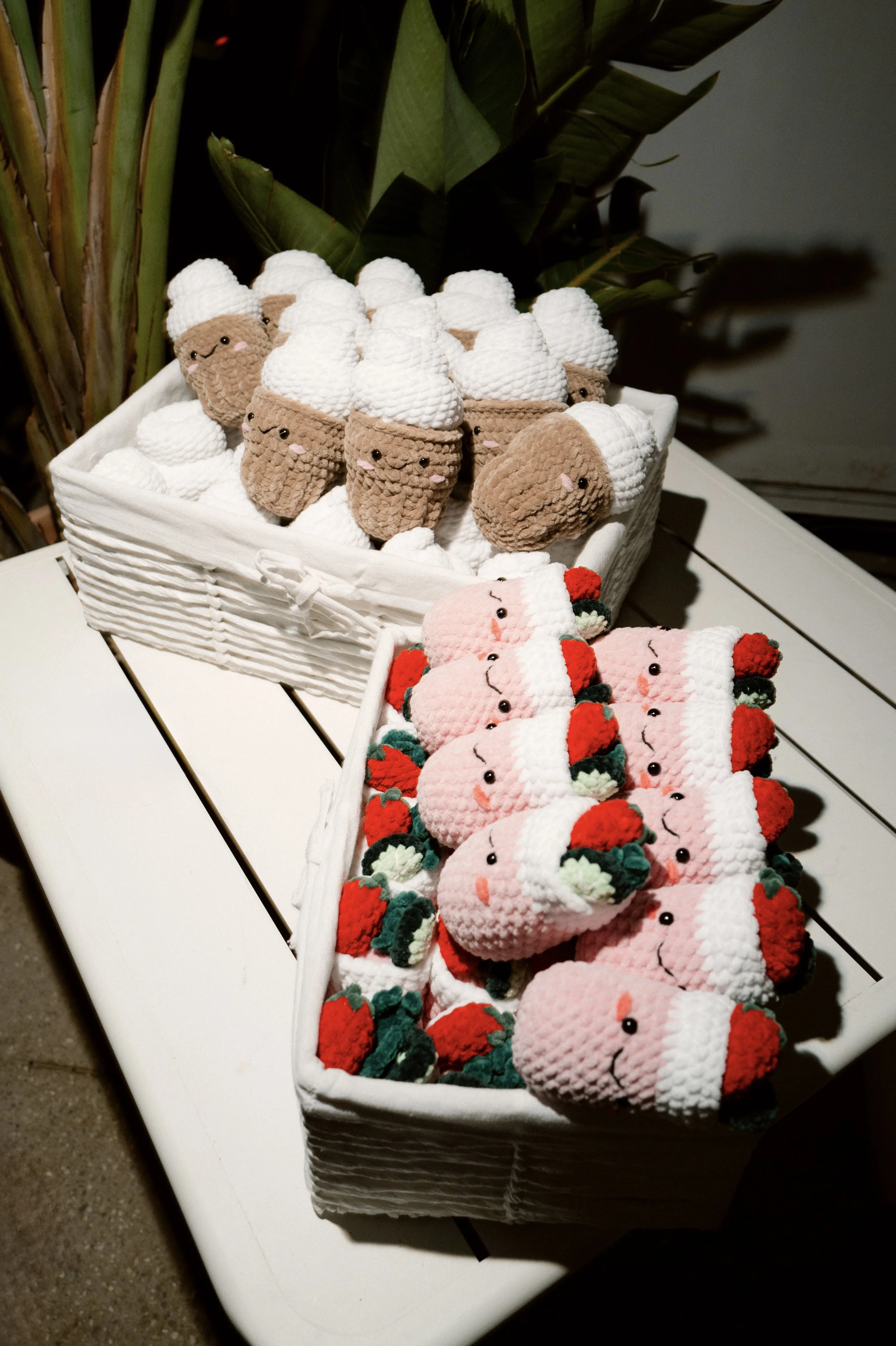 Display of handmade plush toys shaped like ice cream and strawberries with smiling faces in white baskets on a white table, with green plants in the background.