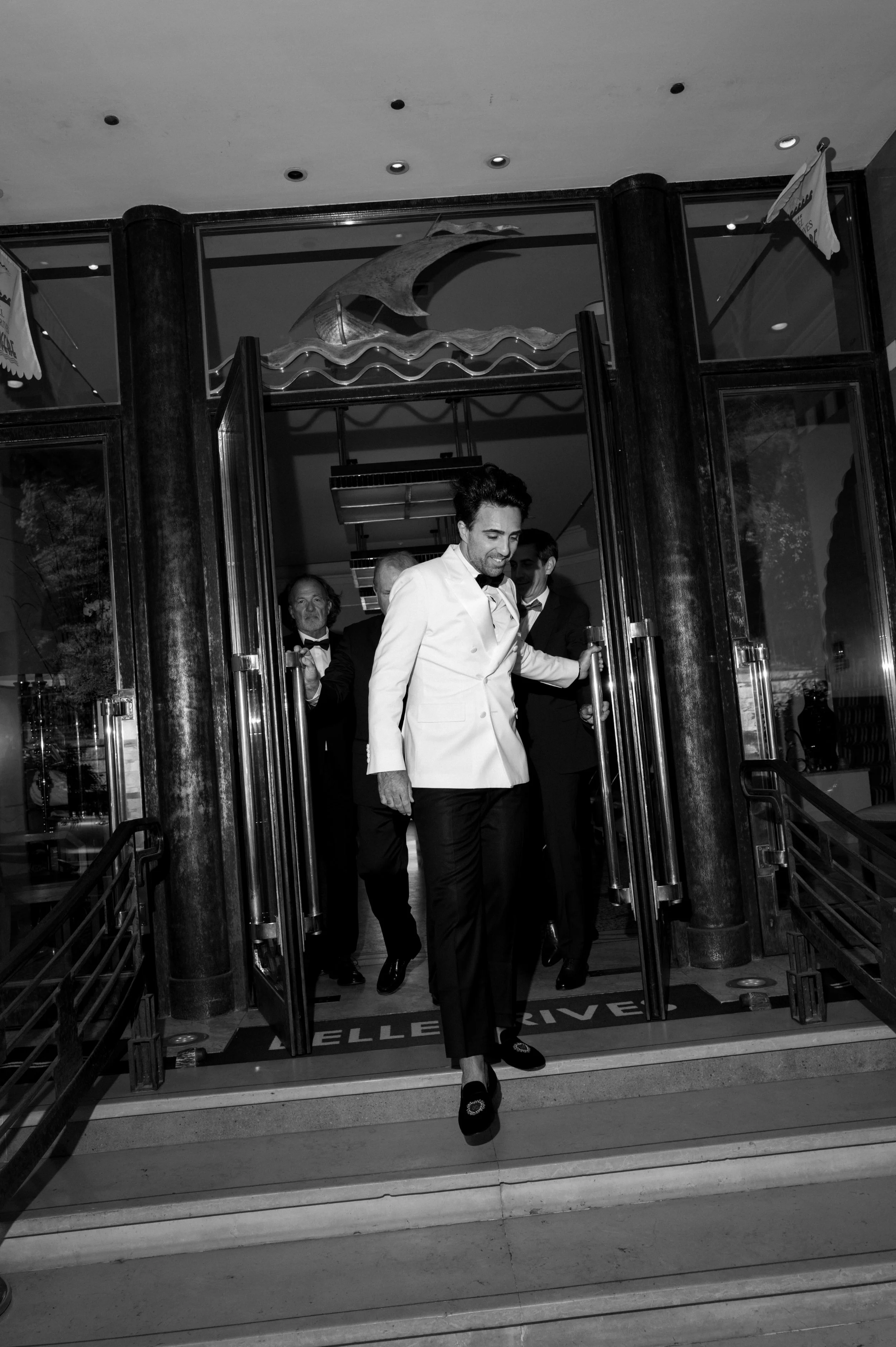 People exiting a restaurant or hotel at night, with a man in front wearing a white jacket and dark pants, smiling as he opens the door, with others following behind.