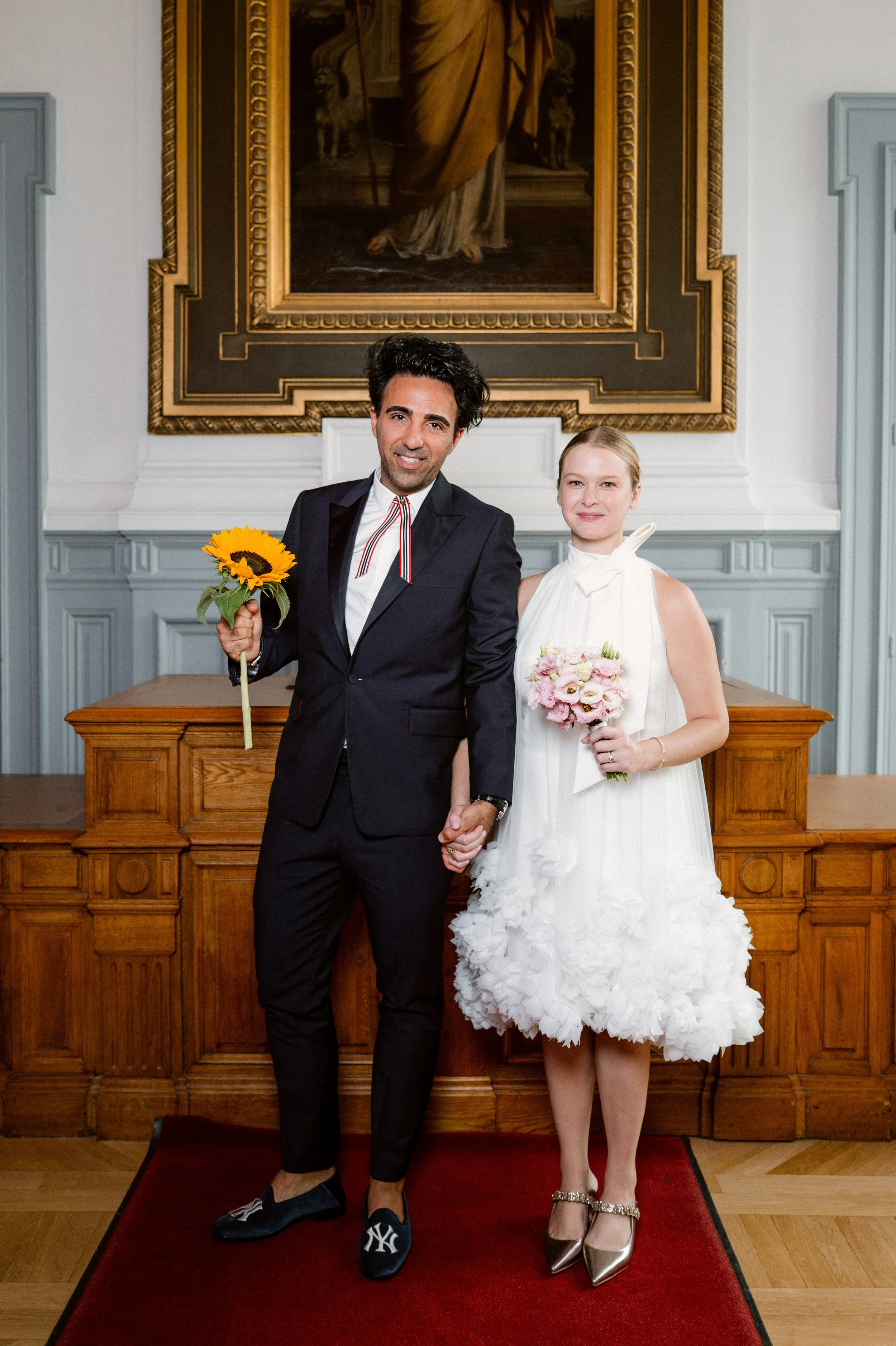 A man and woman holding hands in a formal setting. The man is holding a sunflower, and the woman is holding a pink bouquet. They are standing on a red carpet in front of a wooden table with a large framed painting above them.