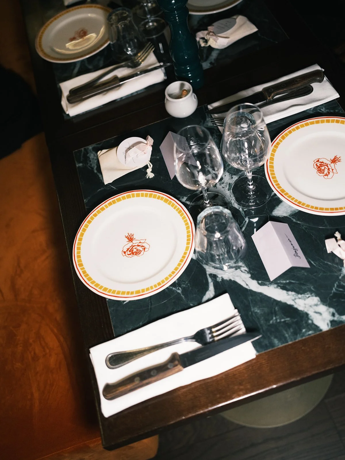 A table set for a meal with two plates with a yellow and red floral design, three wine glasses, silverware wrapped in white napkins, a small white sauce dish with a spoon, and a small white card on a dark green marble surface.