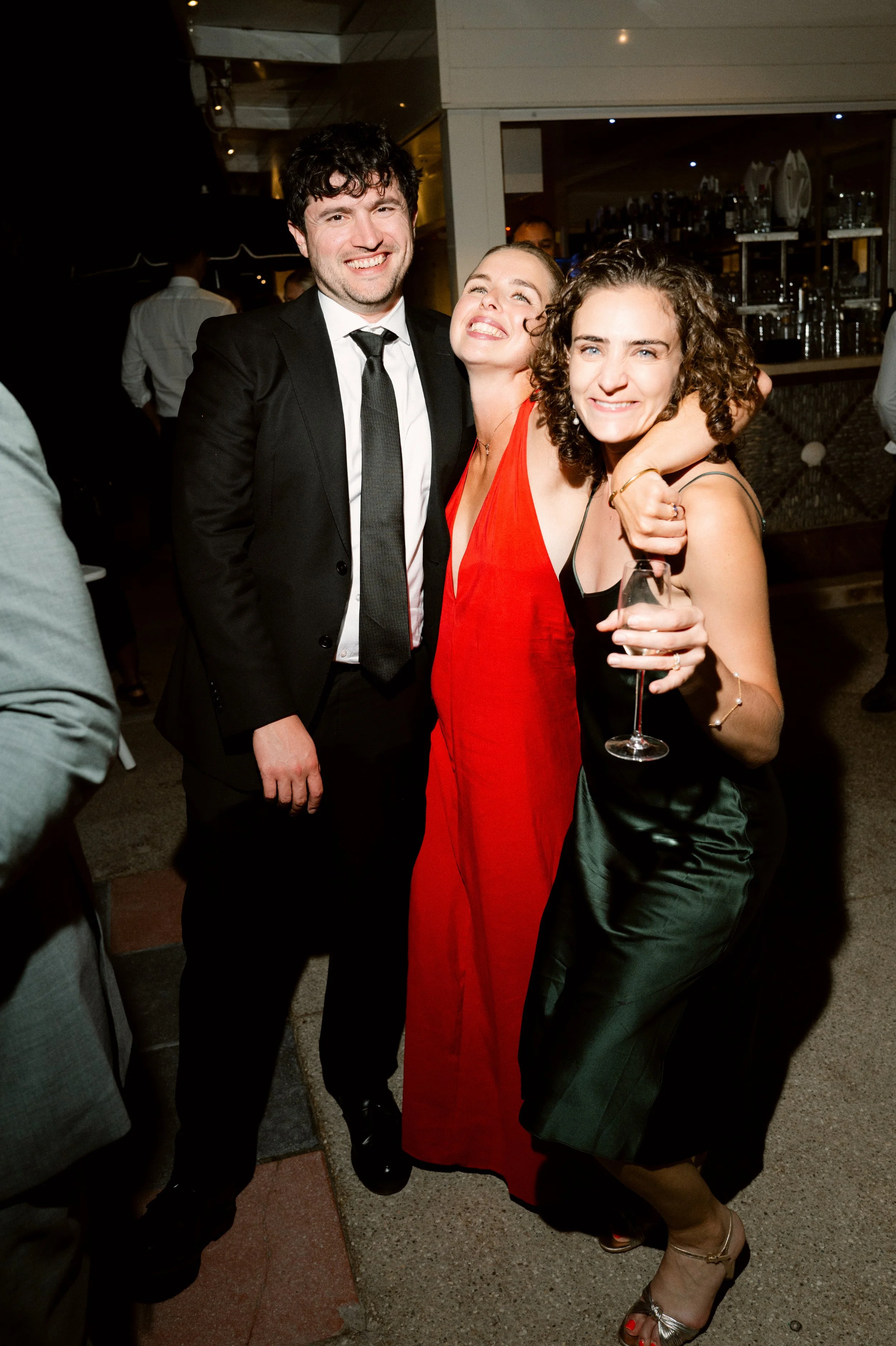 Three friends, two women and one man, enjoying a night out at a bar, smiling, with the woman on the right holding a glass of wine.