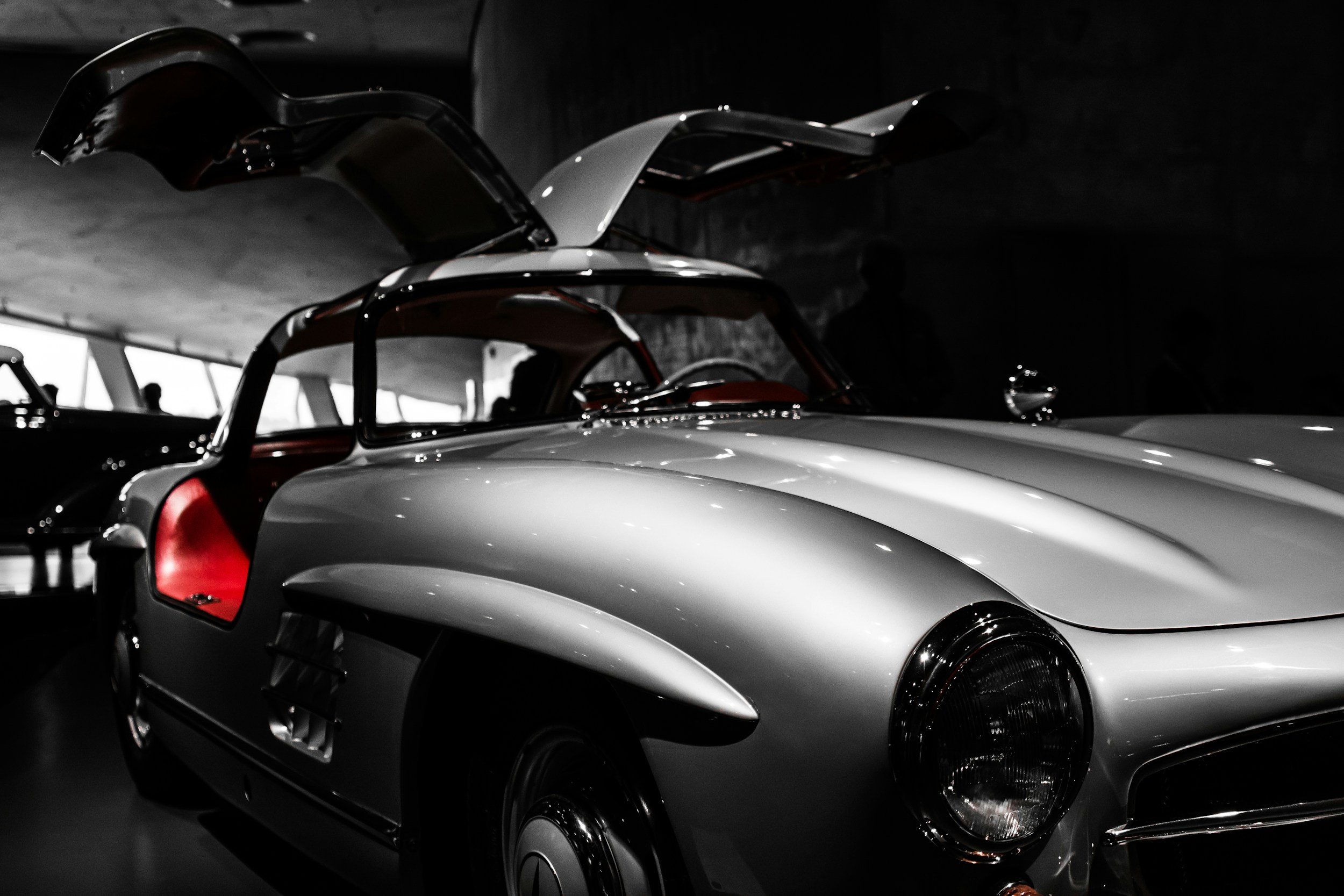 Black and white photograph of a vintage silver sports car with a distinctive curved shape, open gullwing doors revealing a red interior, displayed in a dimly lit museum or showroom.