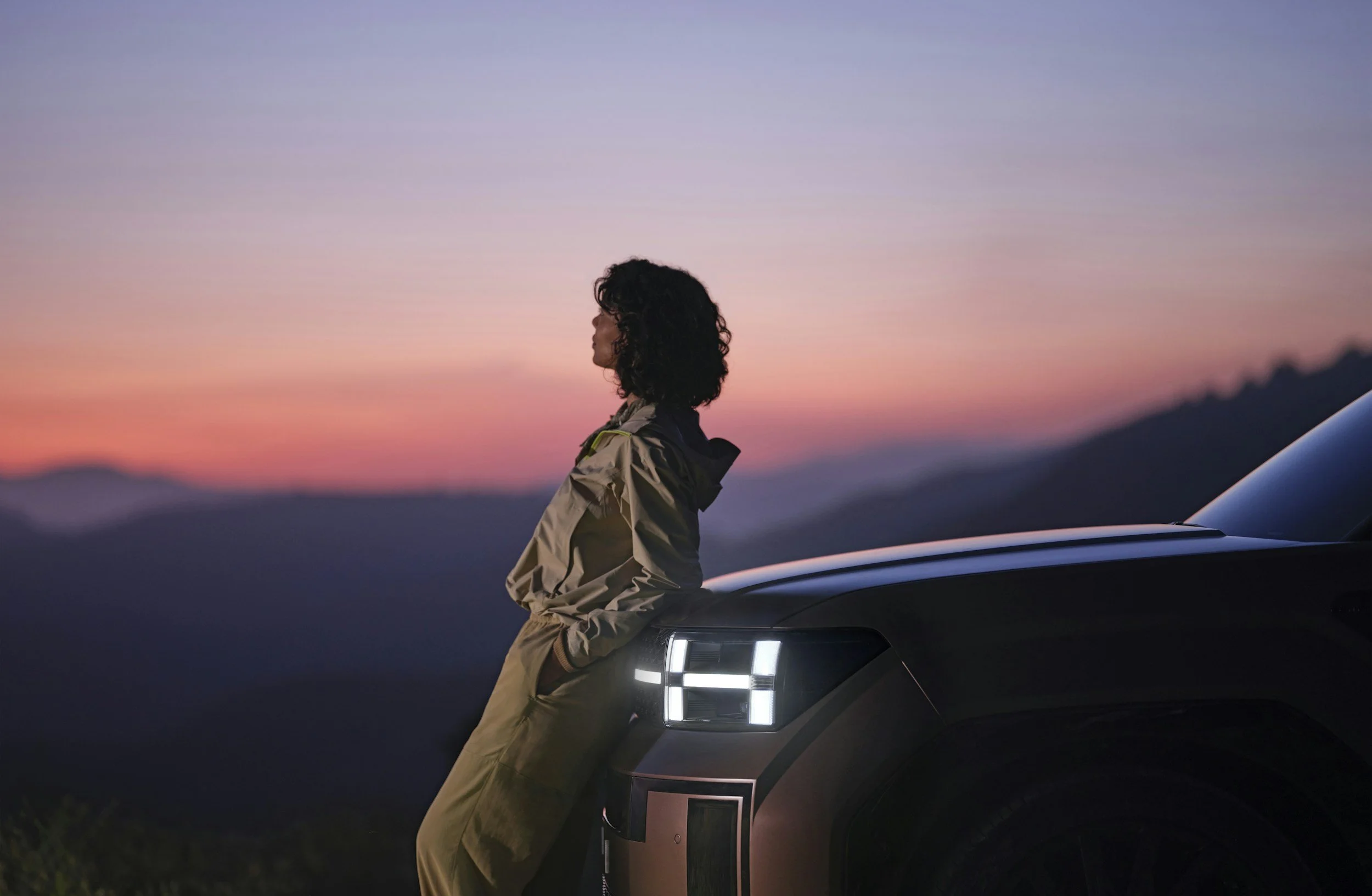 A woman with curly hair leaning against the front of a dark-colored SUV during sunset, with mountains in the background.