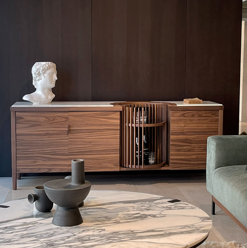 A wooden sideboard with a white bust sculpture on the left, a stack of papers on the right, and a circular shelving unit in the center holding glasses and bottles. In front, there are three ceramic vases on a marble-top table. A green couch is partly visible on the right against a dark wood-paneled wall.