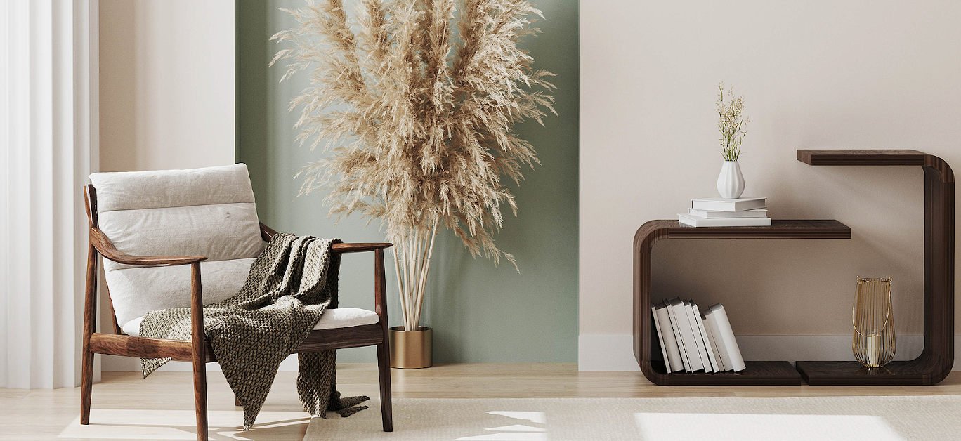 Modern living room corner with a light-colored chair draped with a blanket, a tall pampas grass plant in a gold vase, and a dark wooden bookshelf with books and decorative items, against a two-tone wall and a wood floor.