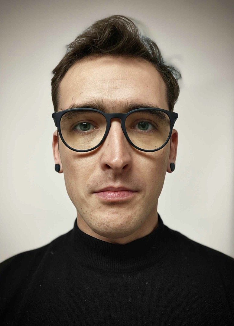 A young man with glasses, black earrings, and a black turtleneck, looking directly at the camera with a neutral expression, against a plain light-colored background.