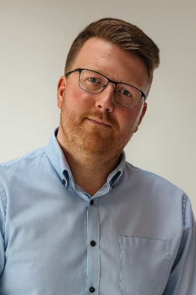 A man with glasses and brown hair wearing a light blue button-down shirt, looking at the camera against a plain background.