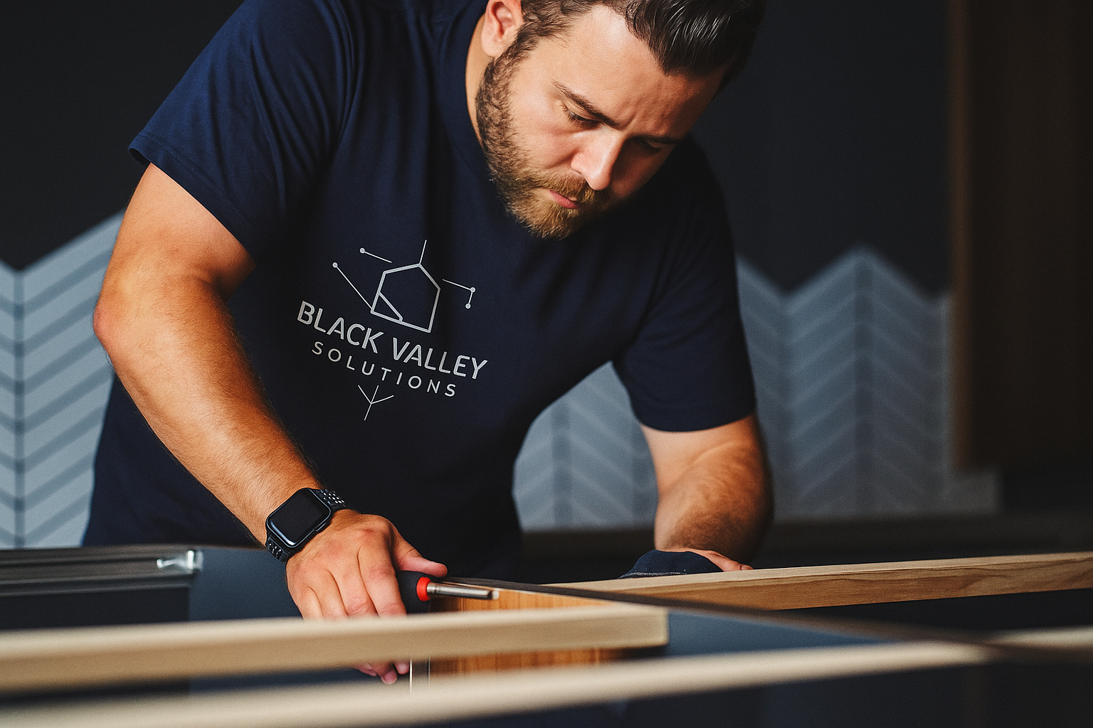 A man wearing a black T-shirt with 'Black Valley Solutions' logo measuring a piece of wood with a square tool in a woodworking shop.